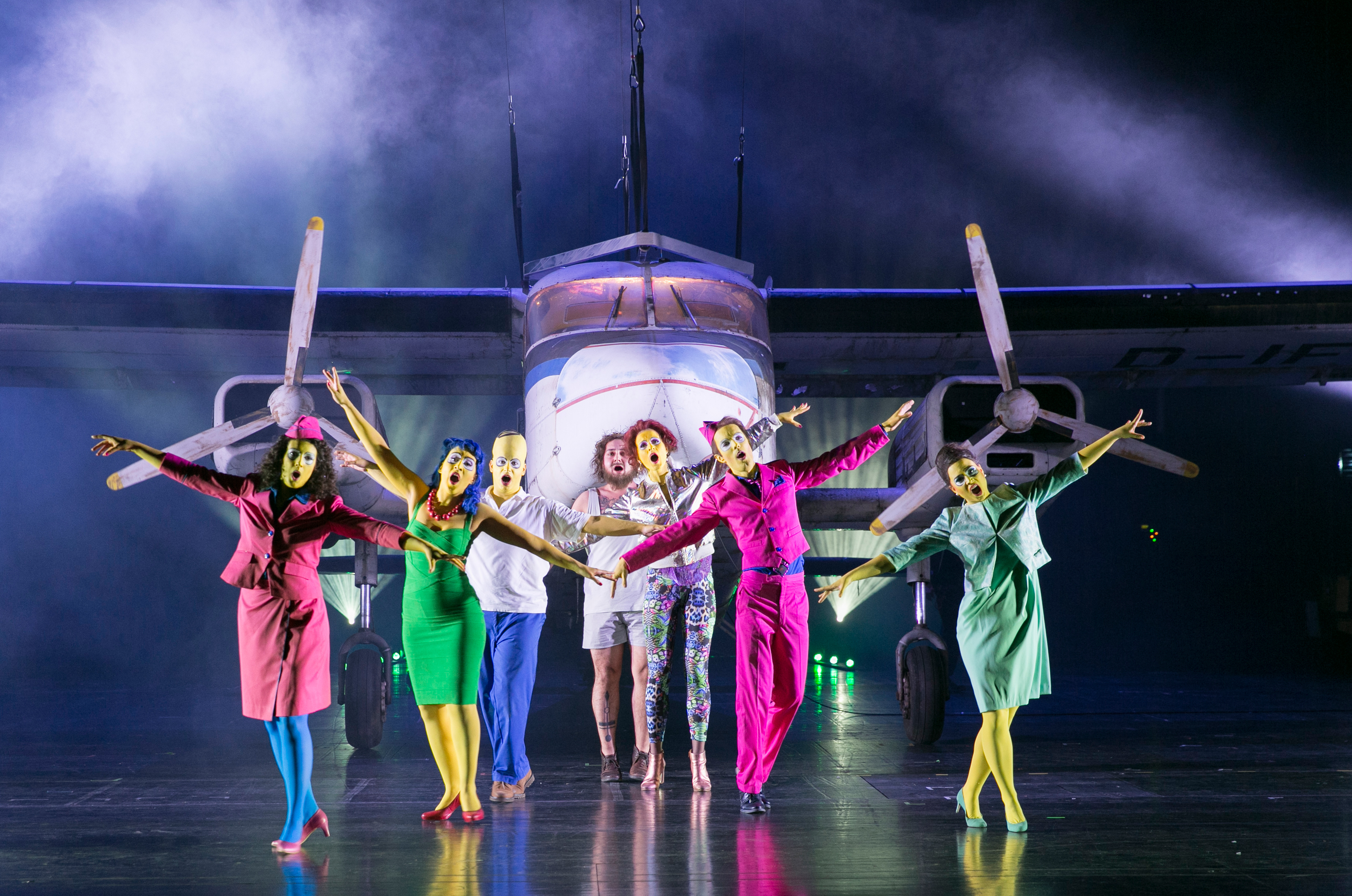 Bunt gekleidete, gelb geschminkte Sänger:innen tanzen auf der Bühne vor einem maßstabsgetreuen Flugzeug