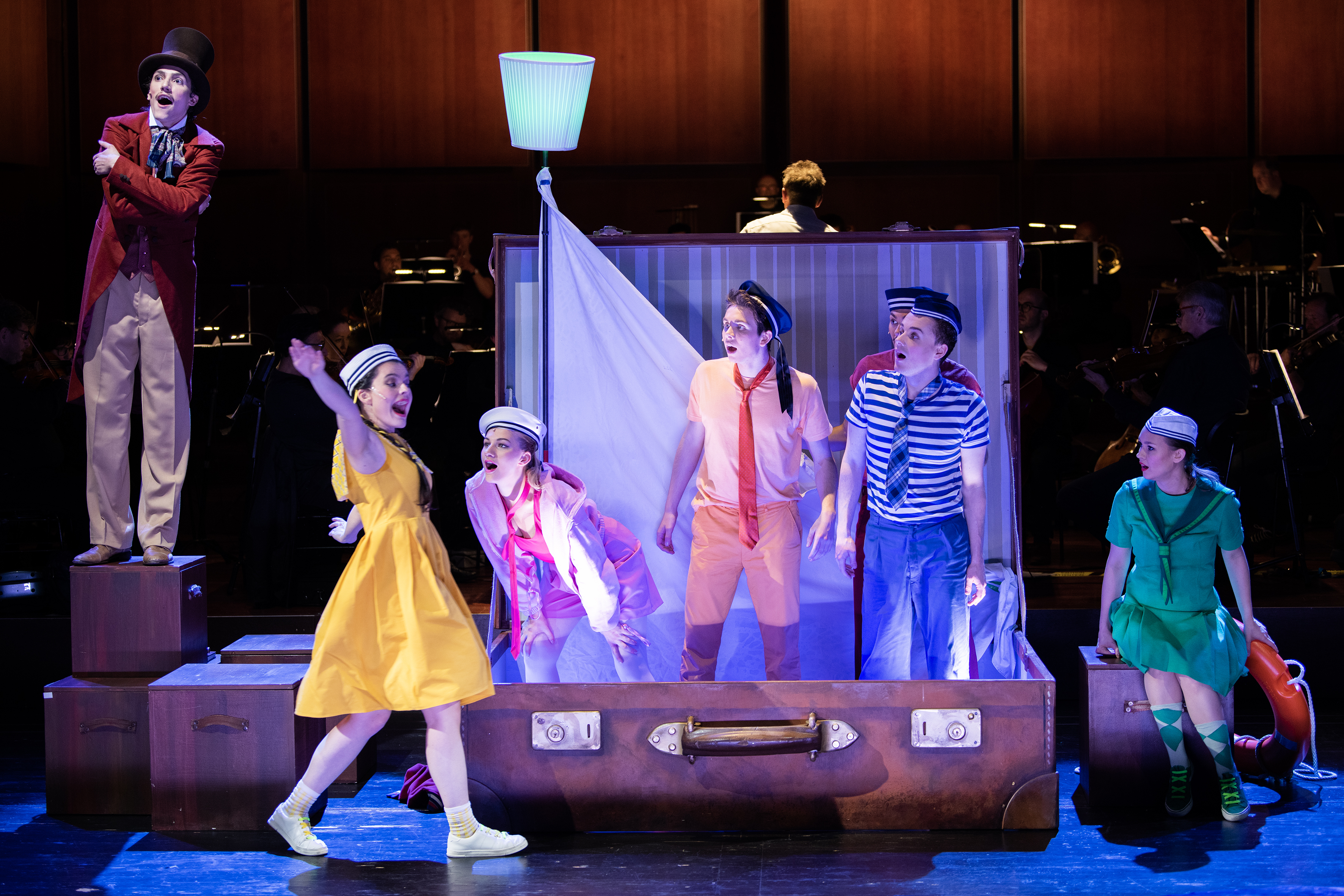 Bunt gekleidete Sänger:innen stehen in einem übergroßen Reisekoffer, links ein Mann in rotem Frack und Zylinder