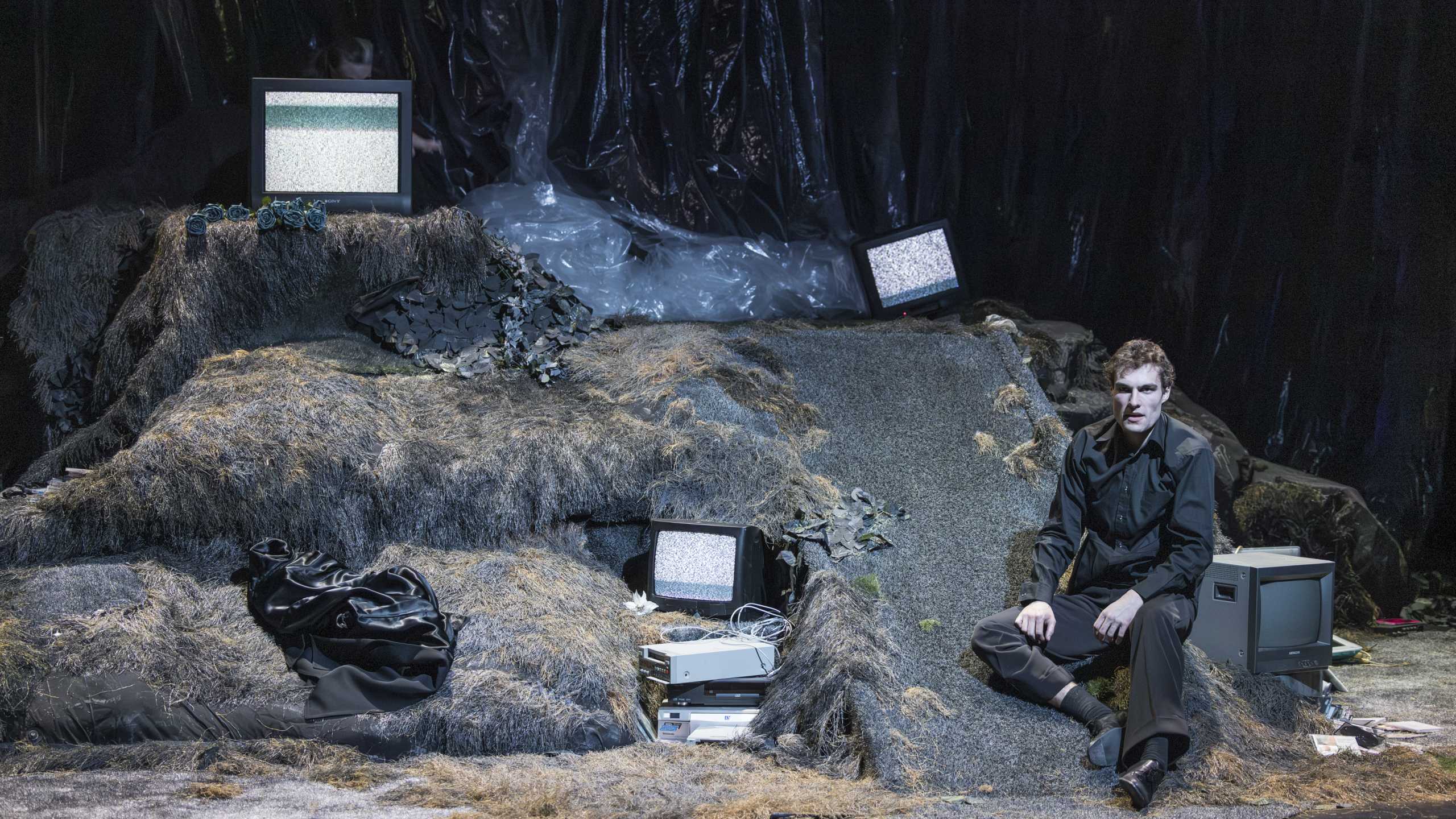 Die Figur Hamlet sitzt auf einem Erdhügel mit alten Modems und Fernsehgeräten. Inszenierung von Ondrej Skrabal im Januar 2026.