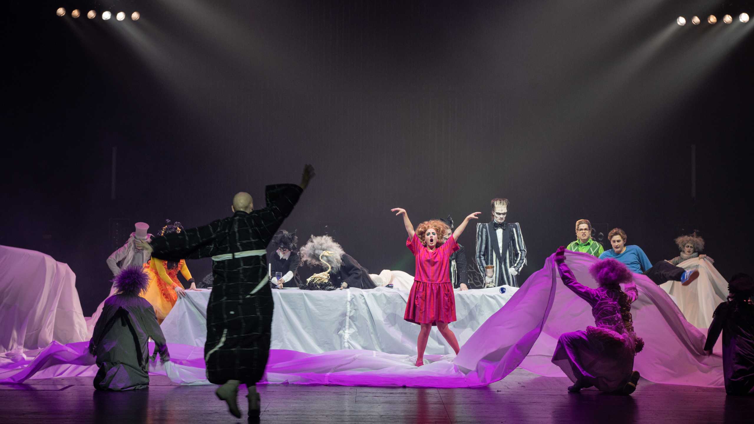 12 kostümierte Personen bewegen sich in alle Richtungen auf einem weißen Tuch, das einen Tisch bedeckt. Szenenbild aus dem Musical The Addams Family.