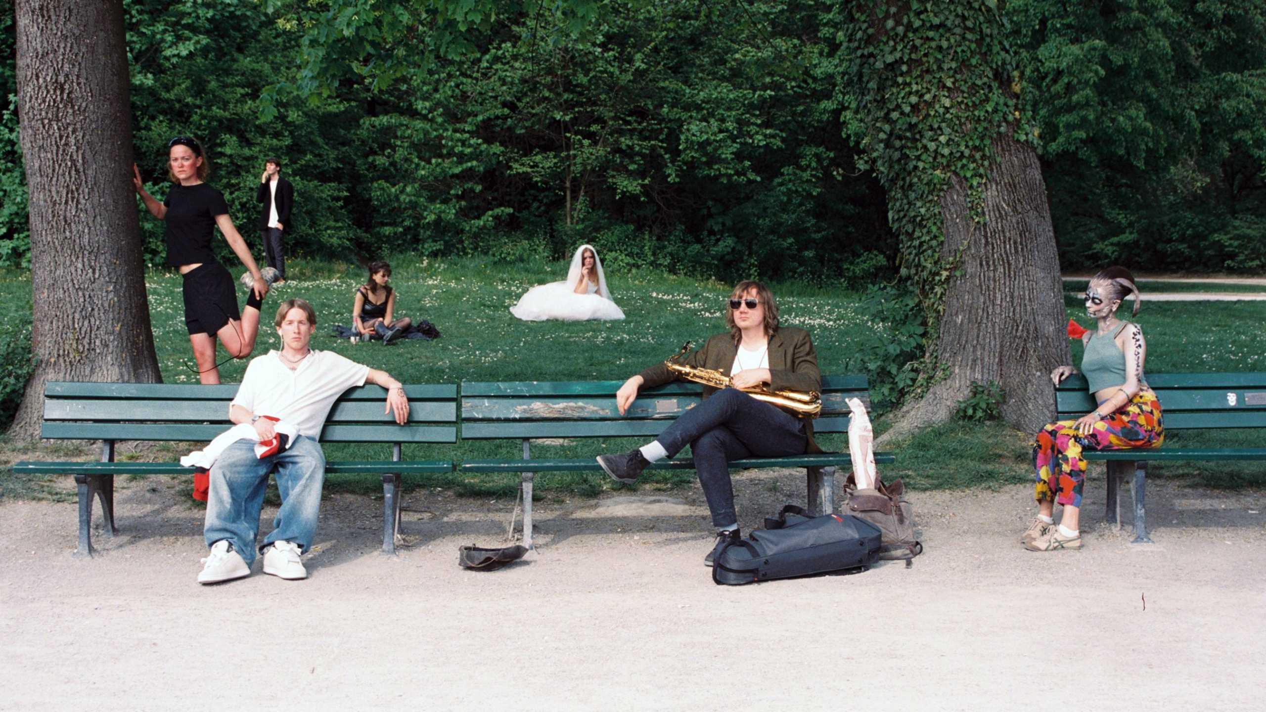 Gruppe von Menschen mit auffälligen Kostümen und Masken sitzt und steht auf und neben Parkbänken unter Bäumen.