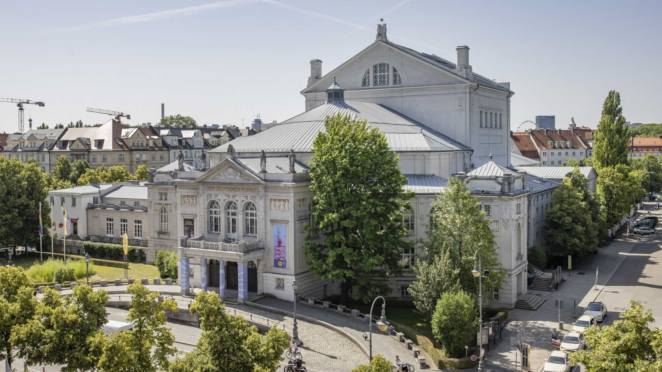 Drohnenaufnahme vom Prinzregententheater bei Sonnenlicht von ca. 75 Meter Entfernung. Grüne Bäume und blauer, diesiger Himmel.
