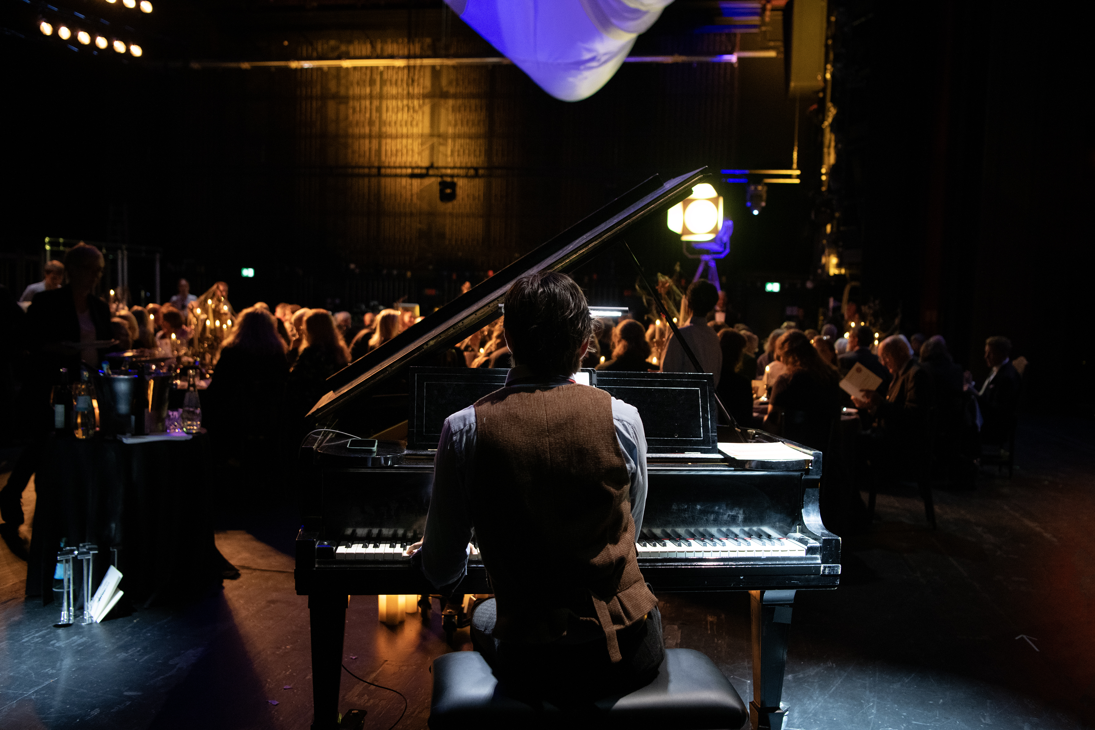 Pianist am Flügel von hinten. Vor sich die Festgesellschaft beim essen, etwa 90 Personen auf einer Bühne. Eiserner Vorhang im Hintergrund