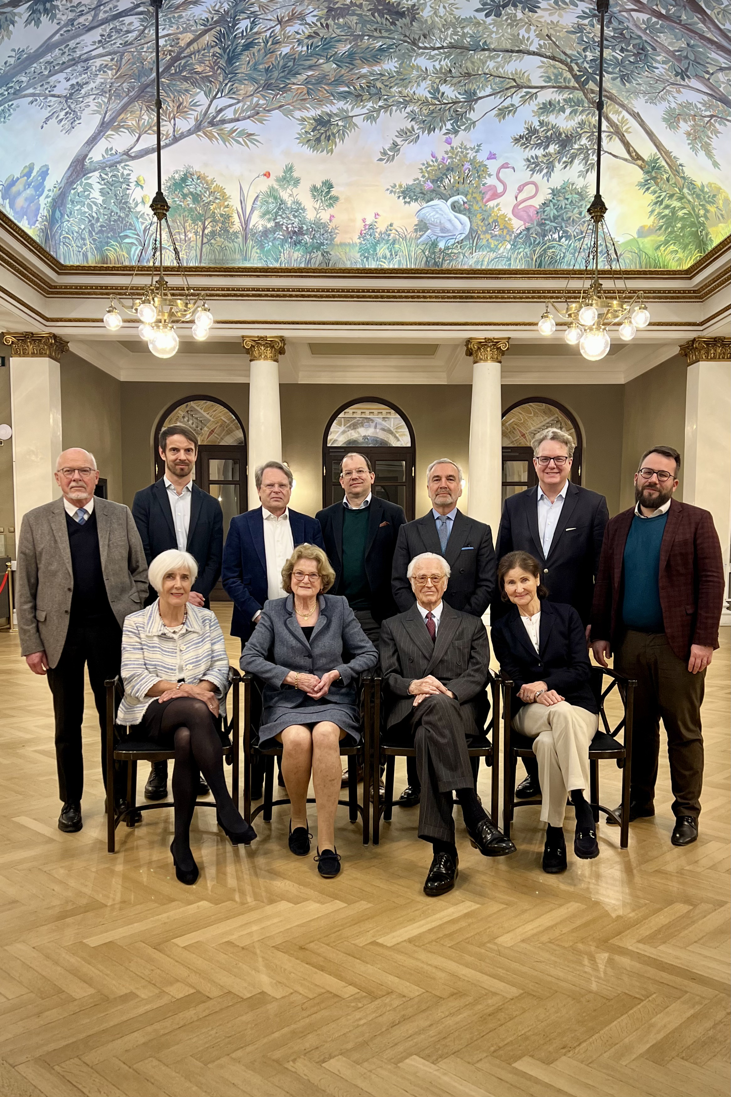 Gruppe von elf Personen in formeller Kleidung im prunkvollen Gartensaal mit bemaltem Deckenfresko im Prinzregententheater München