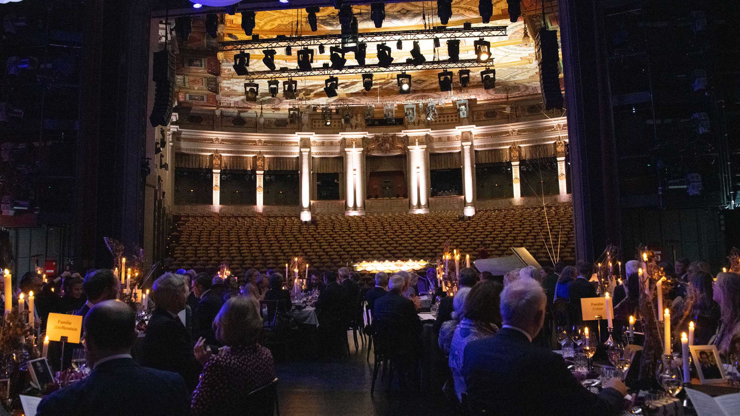 Dinnertische mit sitzenden Menschen in Abendkleidung auf der Bühne mit Blick auf den erleuchteten, leeren Zuschauerraum des Prinzregententheaters