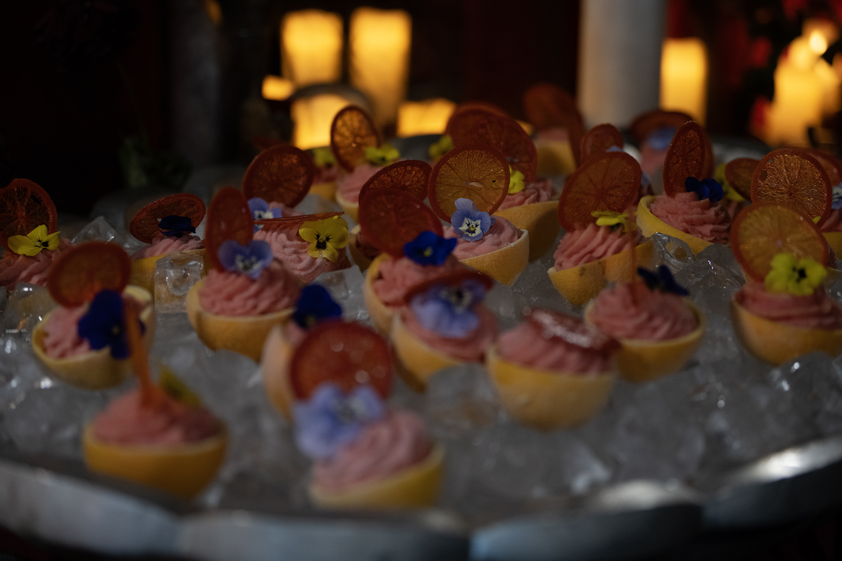 Mehrere kleine Dessertgläser mit rosa Creme, garniert mit getrockneten Orangenscheiben und essbaren Blüten, auf Eiswürfeln arrangiert