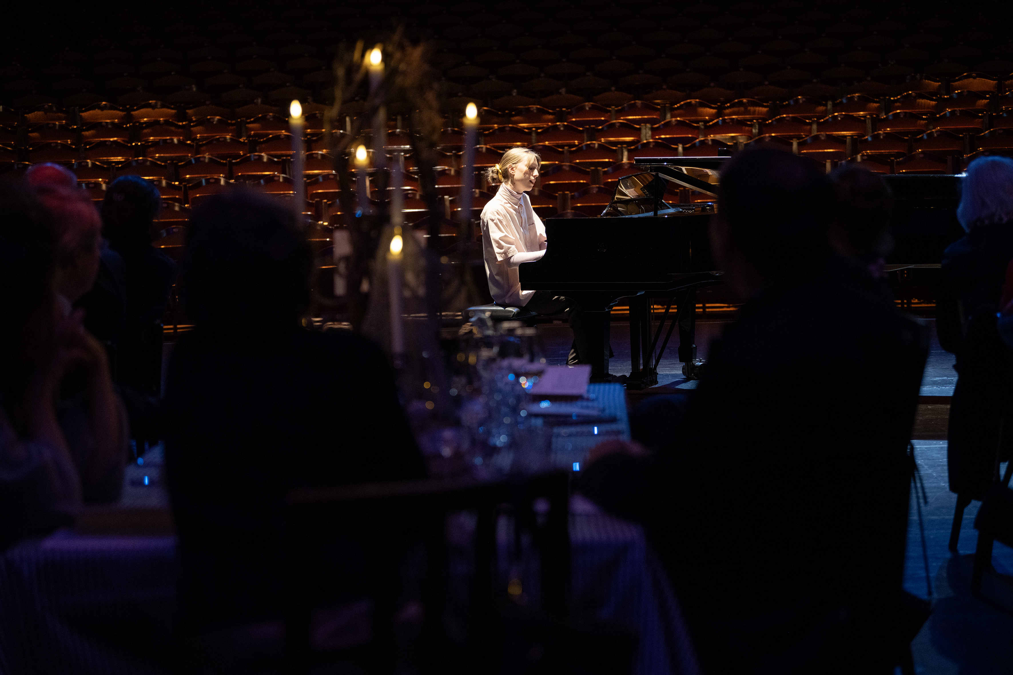 Frau spielt Klavier auf Bühne vor Publikum in dunklem Saal mit Kerzenlicht