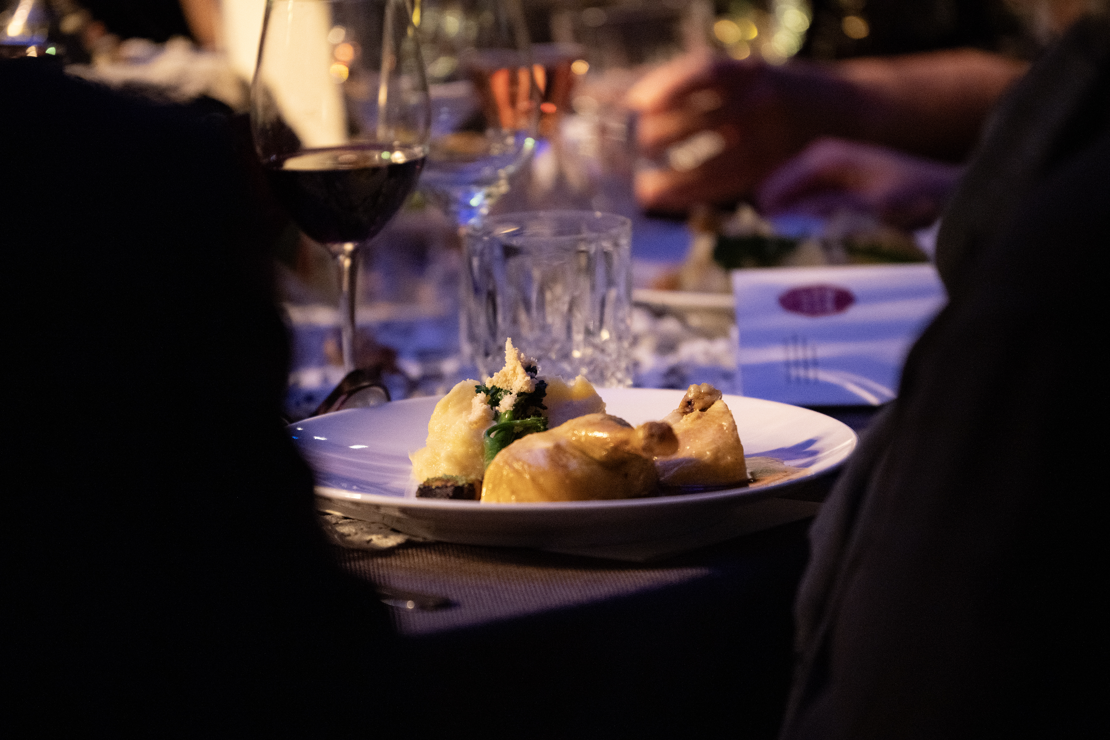 Teller mit einer Portion Essen und einem Glas Rotwein auf einem gedeckten Tisch, umgeben von Personen in dunkler Kleidung