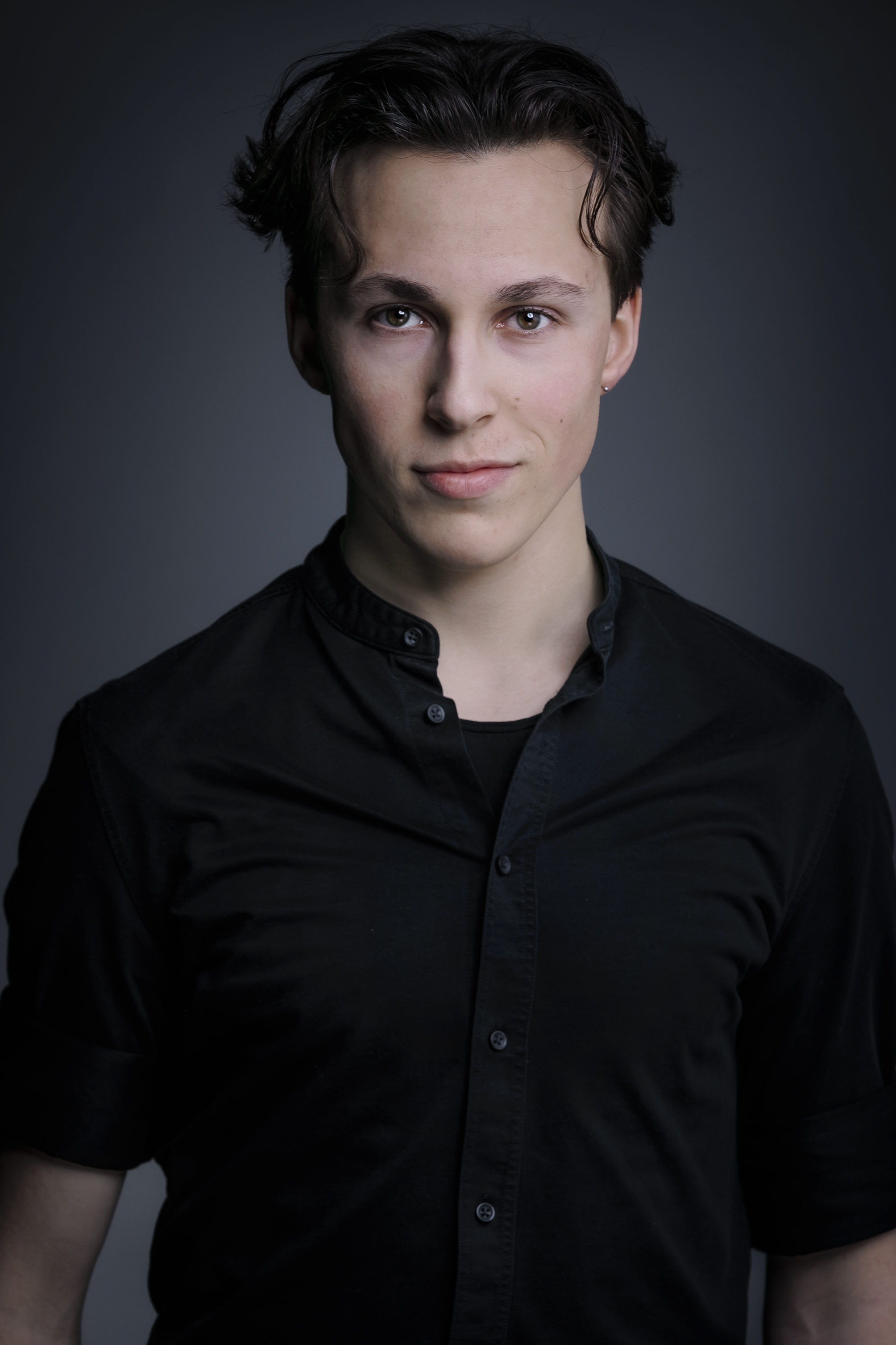 Portraitfoto von Lukas Lommer: Junger Mann mit dunklem, gewelltem Haar, der ein schwarzes Hemd mit Stehkragen trägt und direkt in die Kamera blickt