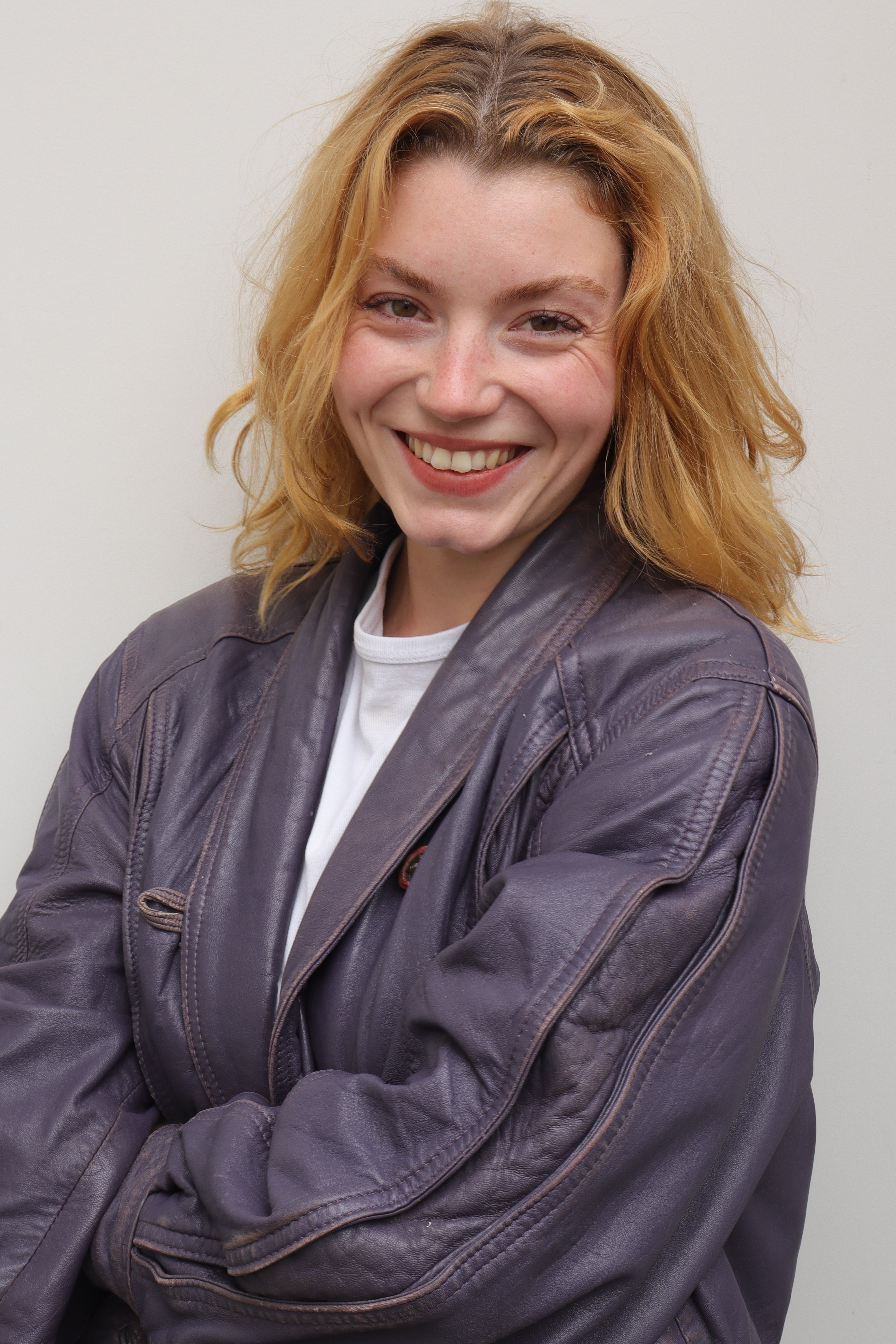 Portraitfoto von Clara Auer: Lächelnde Frau mit lockigem, schulterlangem Haar trägt weiße Bluse und dunkelgraue Lederjacke und hat die Arme verschränkt