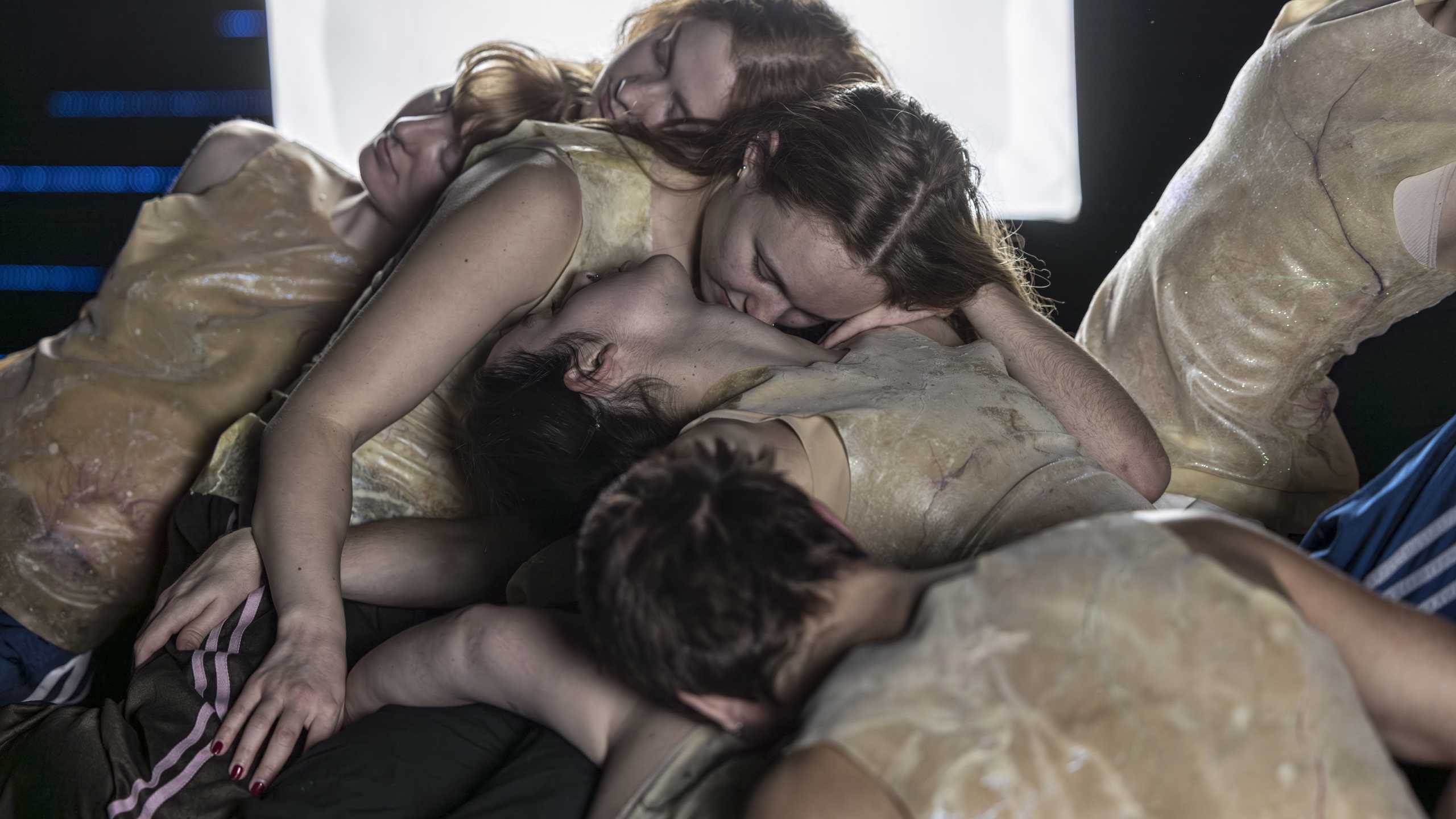 Produktionsbild Living it up way down, ein Knäuel aus circa sieben Tänzer:innen in Kunststoffkostümen in einem stilisierten Bunker