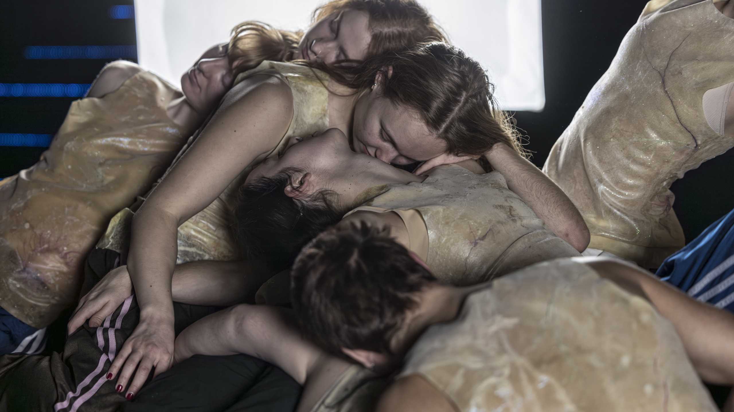 Produktionsbild Living it up way down, ein Knäuel aus circa sieben Tänzer:innen in Kunststoffkostümen in einem stilisierten Bunker