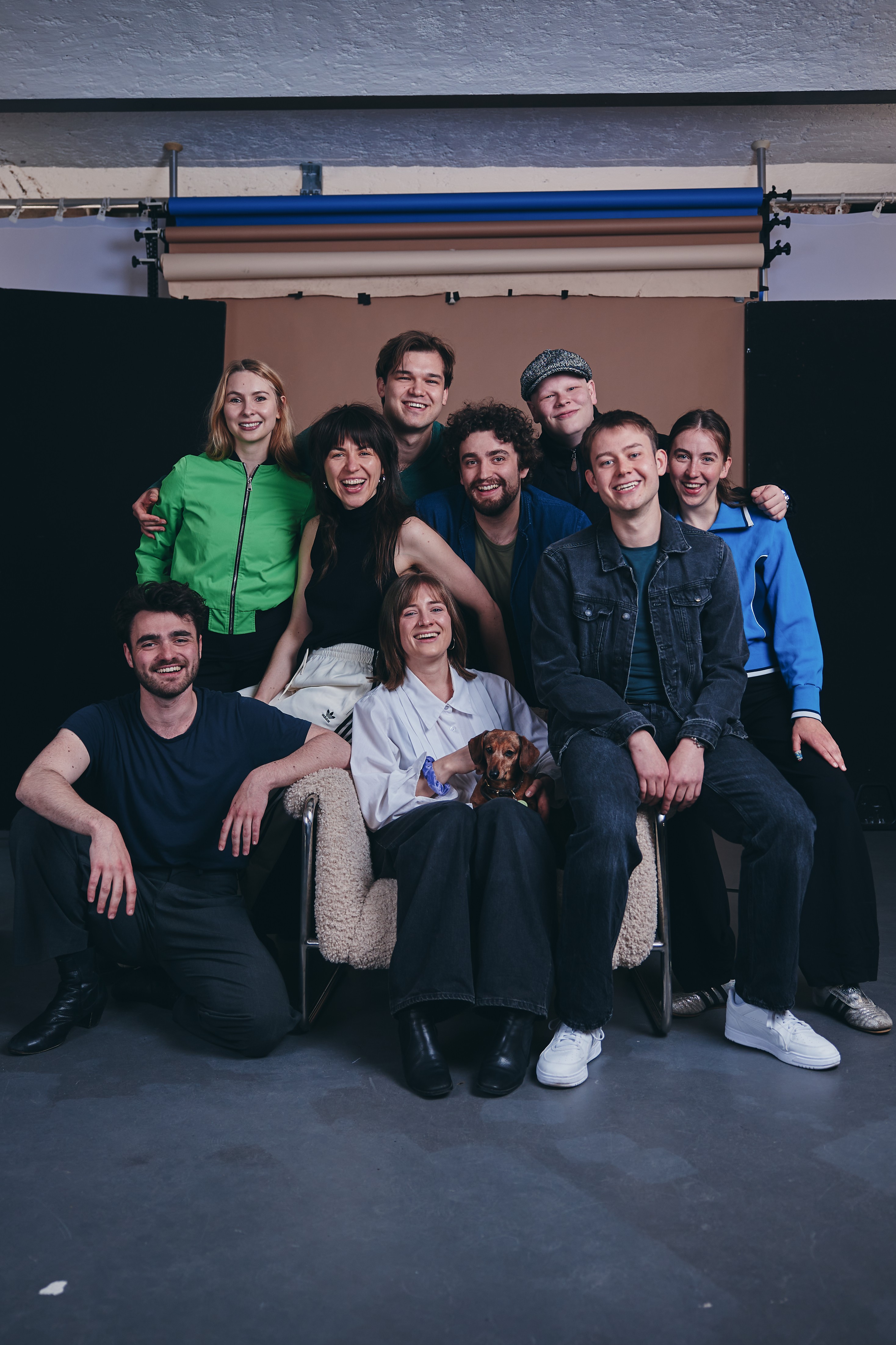 Die neun Absolvent:innen 2027 und ein kleiner Hund sitzen und stehen eng zusammen vor einem Fotohintergrund in einem Studio, von links oben nach unten rechts: Hannah Moreth, Lisa Mader, John Vincent Ragner, Florian Lange, Hannes Tillian, Alexander Schmidt, Frieda Lüttringhaus, Finn-Morten Schuy, Daria Welsch