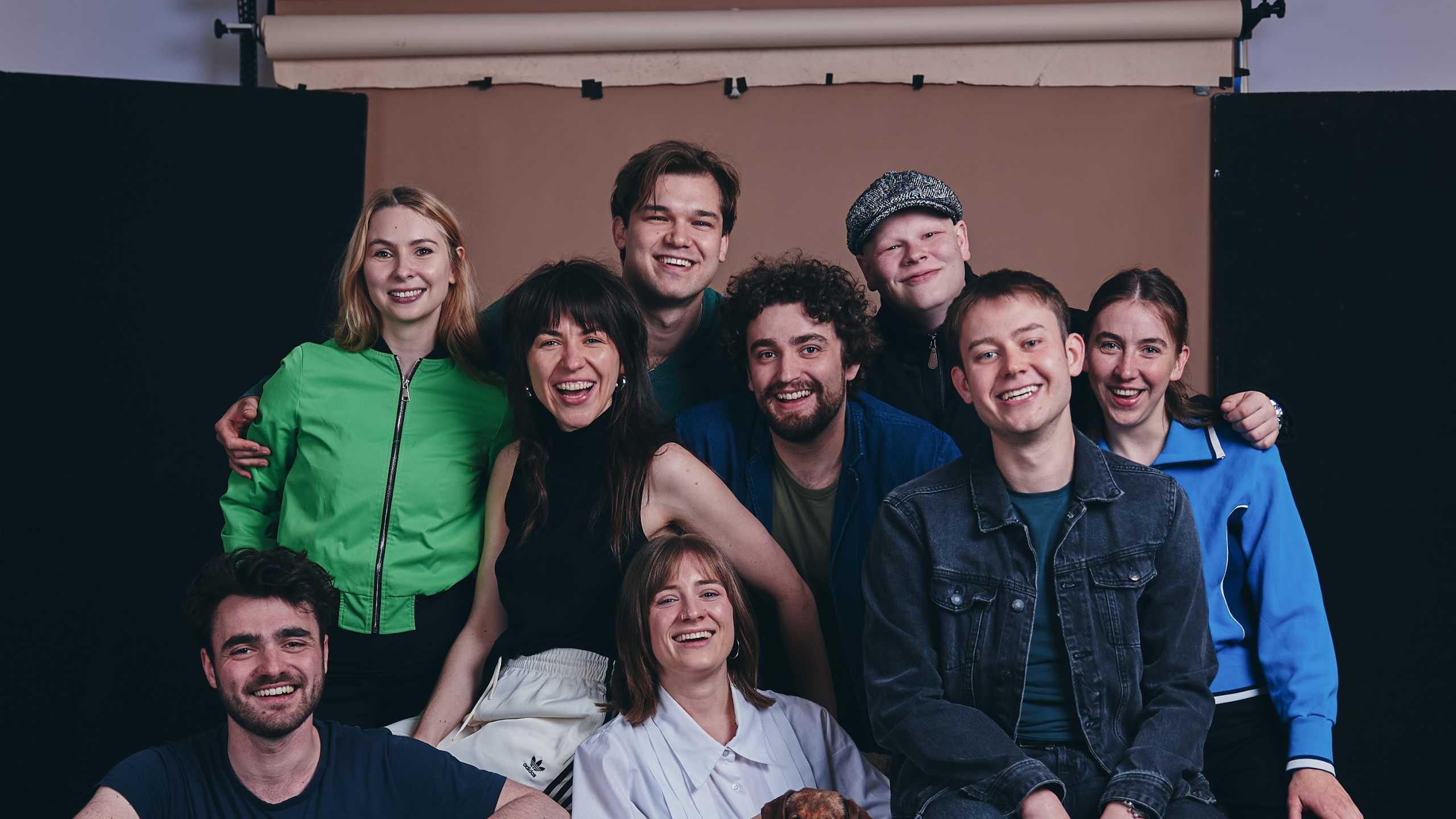 Die neun Absolvent:innen 2027 und ein kleiner Hund sitzen und stehen eng zusammen vor einem Fotohintergrund in einem Studio, von links oben nach unten rechts: Hannah Moreth, Lisa Mader, John Vincent Ragner, Florian Lange, Hannes Tillian, Alexander Schmidt, Frieda Lüttringhaus, Finn-Morten Schuy, Daria Welsch