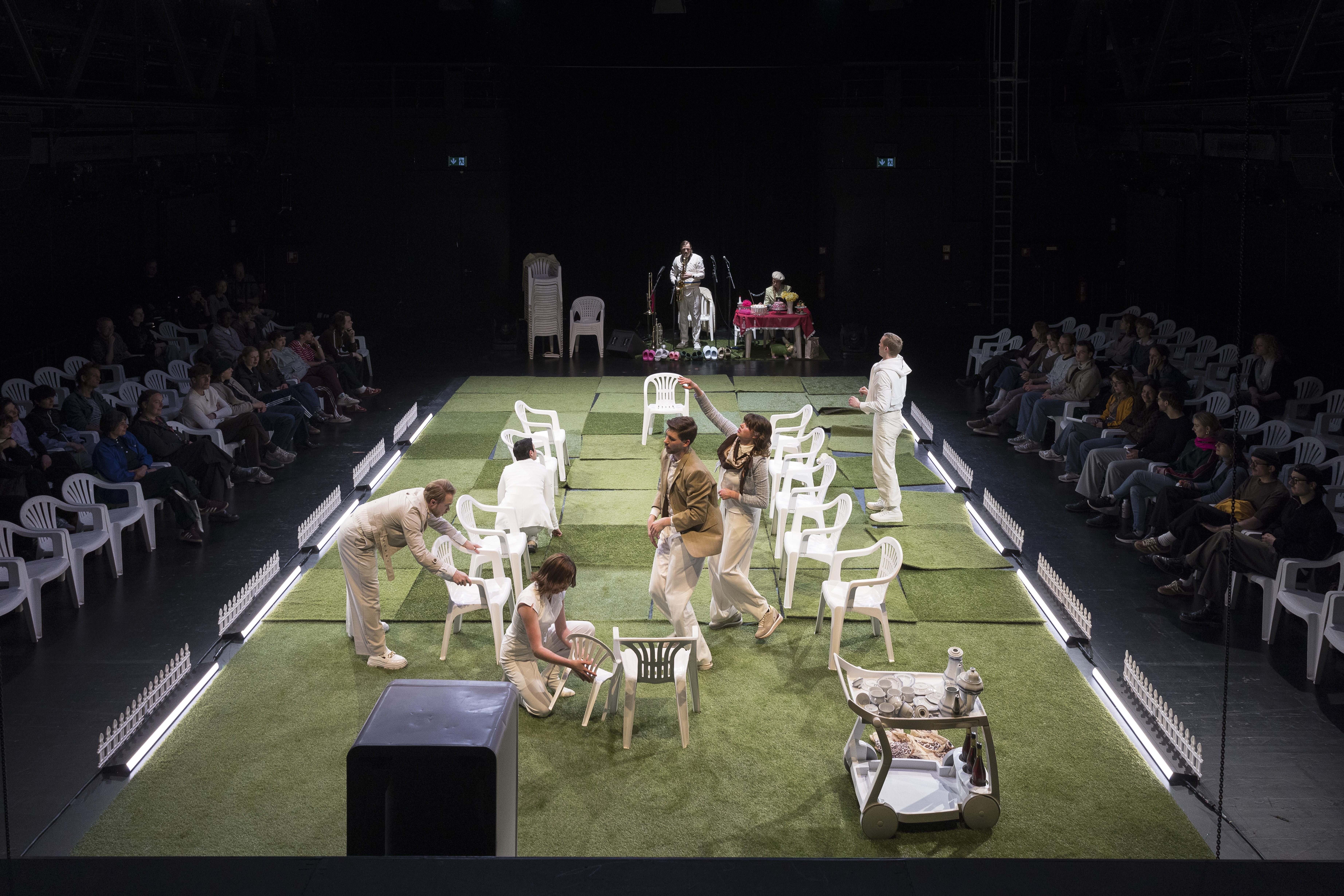 Bühnenbild mit Schauspielern auf grünem Boden und weißen Plastikstühlen, Publikum sitzt links und rechts
