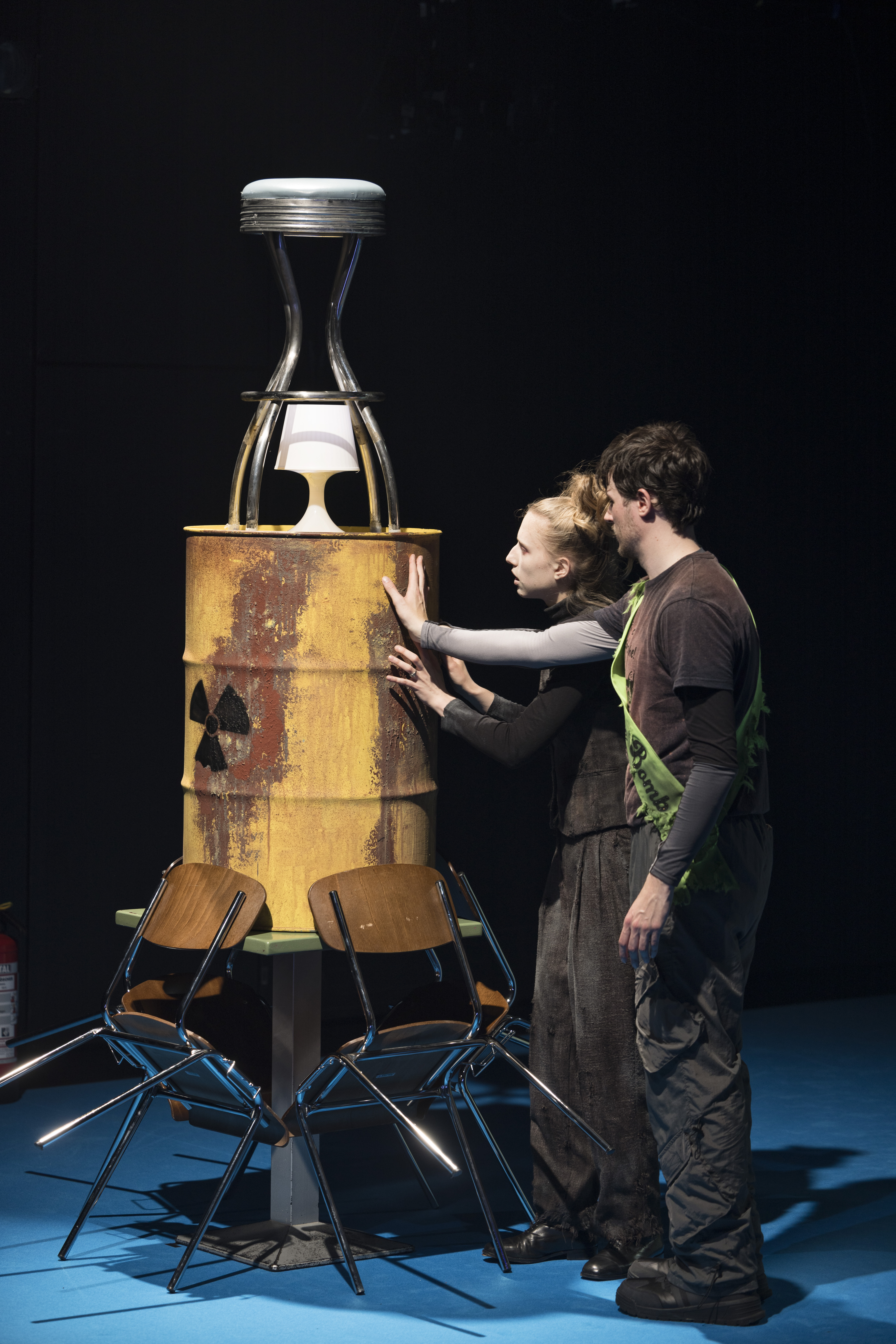 Szenenfoto: Zwei Personen (Rebekka Ziemer und Samuel Spieß) stehen neben einem rostigen Fass mit Strahlensymbol, auf dem eine Tischlampe und ein Barhocker stehen, umgeben von zwei Stühlen.