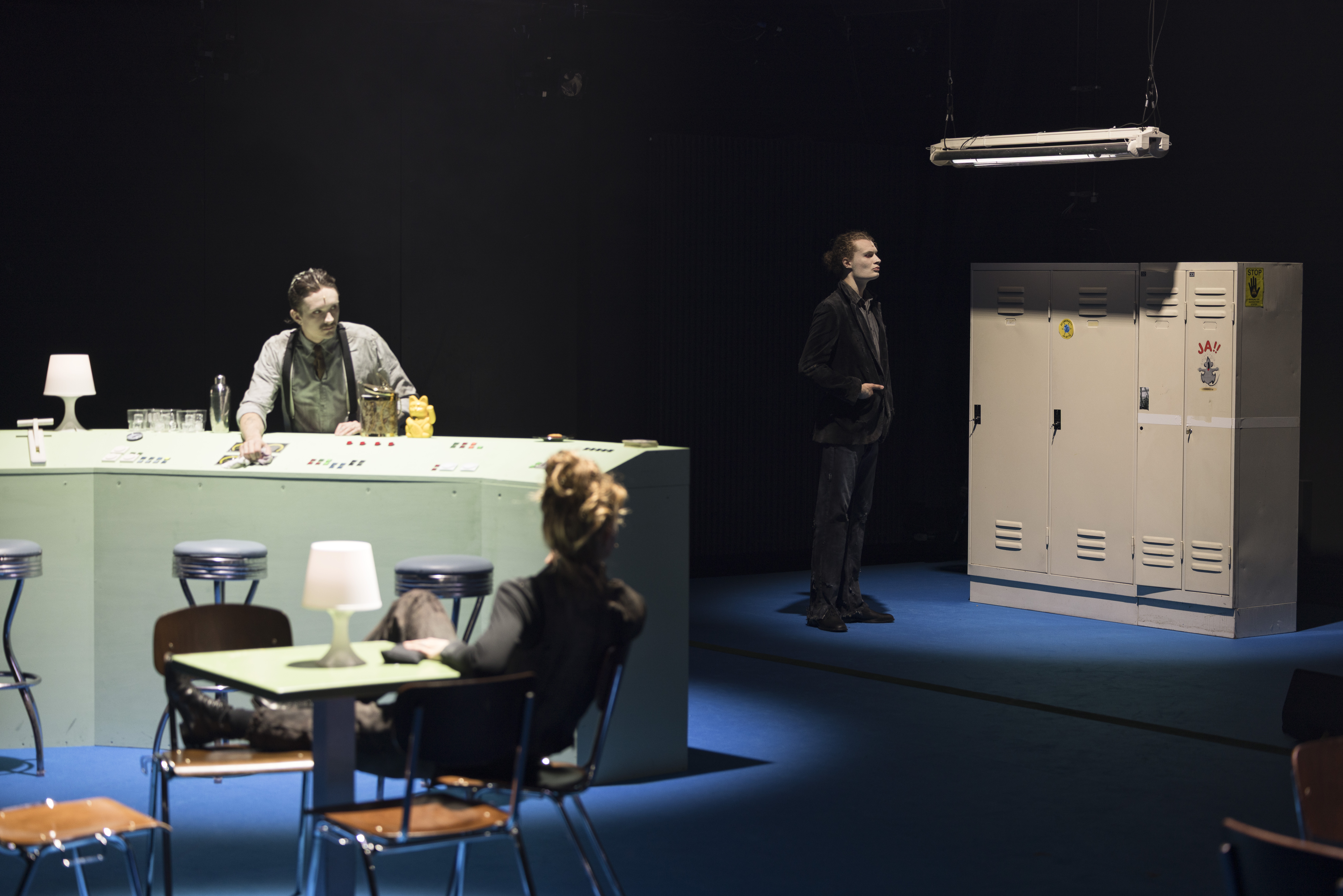 Szenenfoto: Ein Mann (Floyd Clemens) hinter einem Tresen, einer Frau (Rebekka Ziemer) an einem Restauranttisch sowie einem Mann (Bjarne Rentz) vor Schließfächern unter einer Hängelampe mit Neonröhre