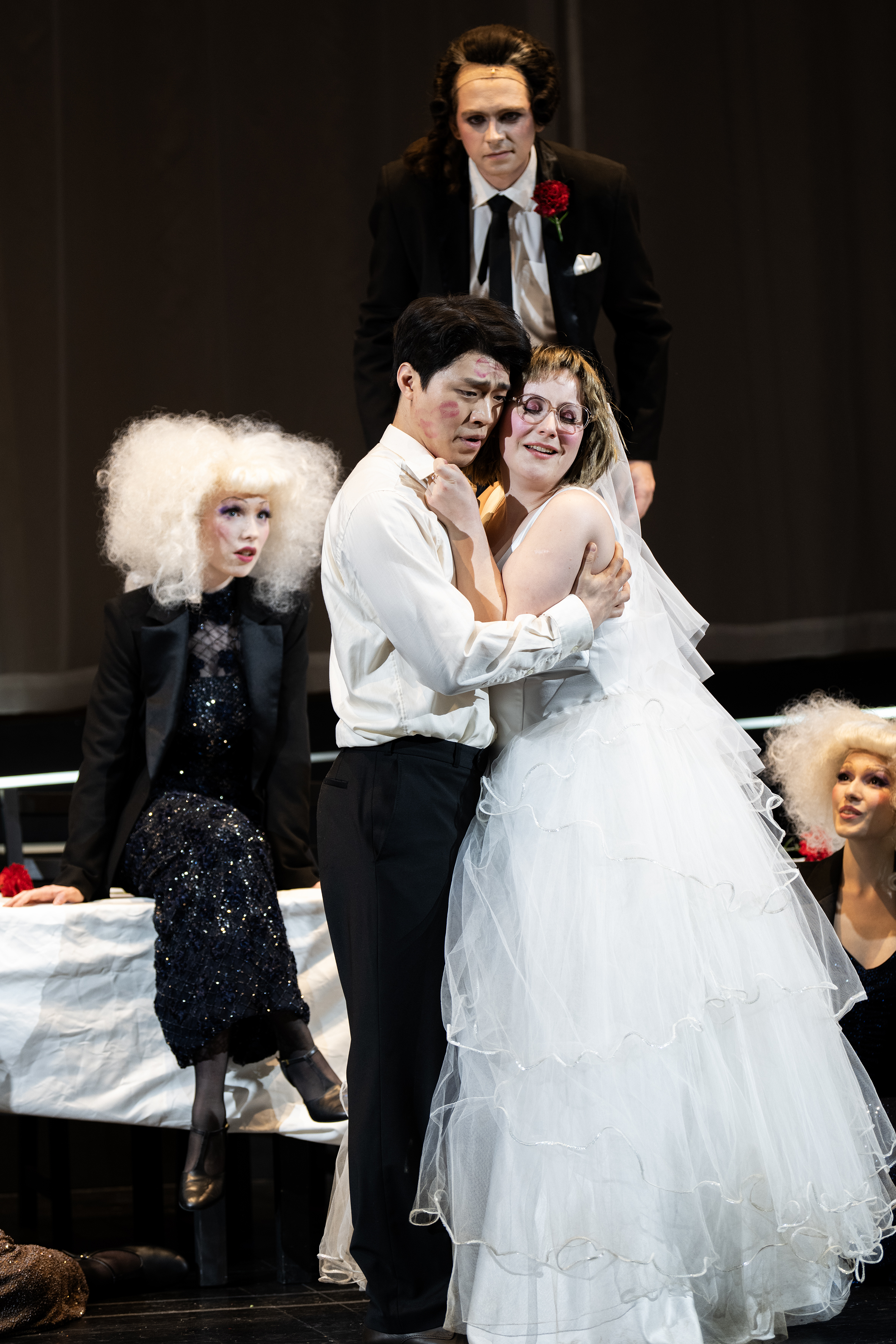 Ein Mann im Anzug und eine Frau im Brautkleid liegen sich in den Armen, um sie herum drei weitere Personen mit neutralem Gesichtsausdruck