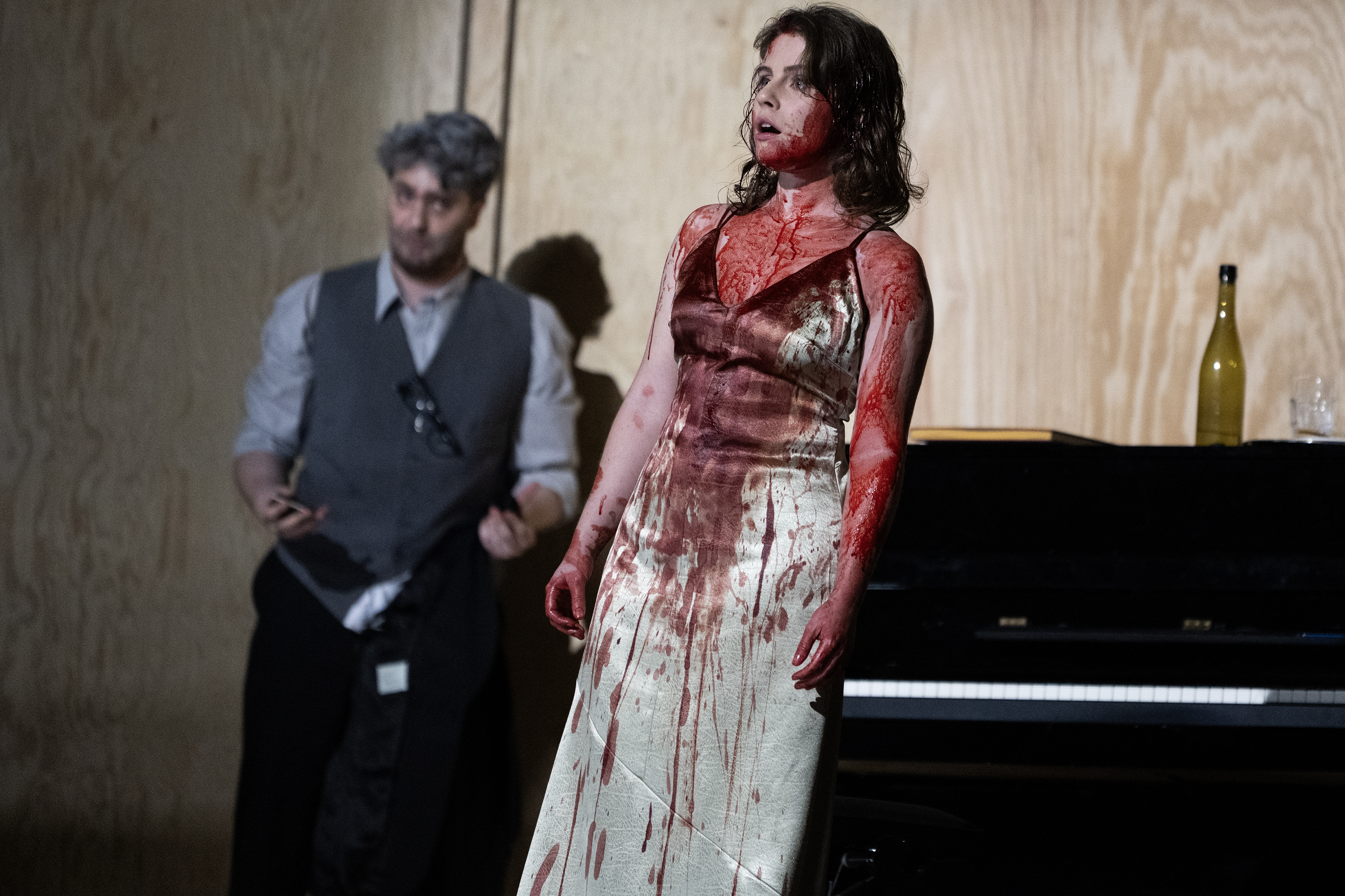Eine Frau mit blutverschmiertem Oberkörper und Kleid, im Hintergrund ein zu ihr blickender Mann und ein Klavier