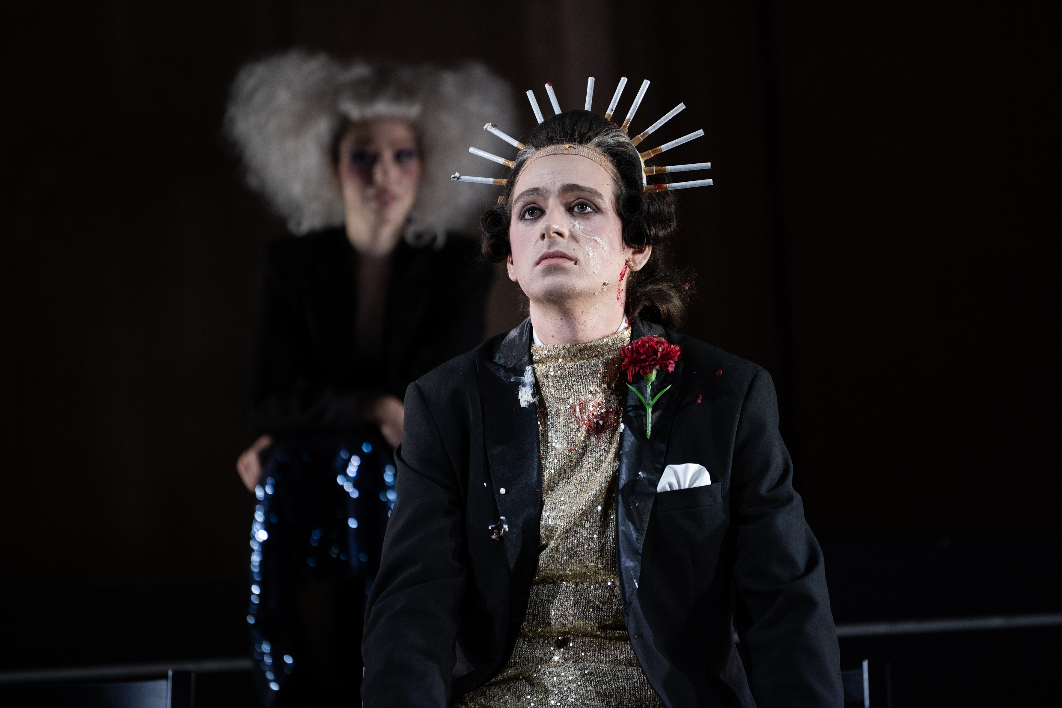 Ein Mann mit roter Rose im Knopfloch seines Jacketts und einer Kopfbedeckung aus Zigaretten, die einen Halbkreis um seinen Kopf bilden