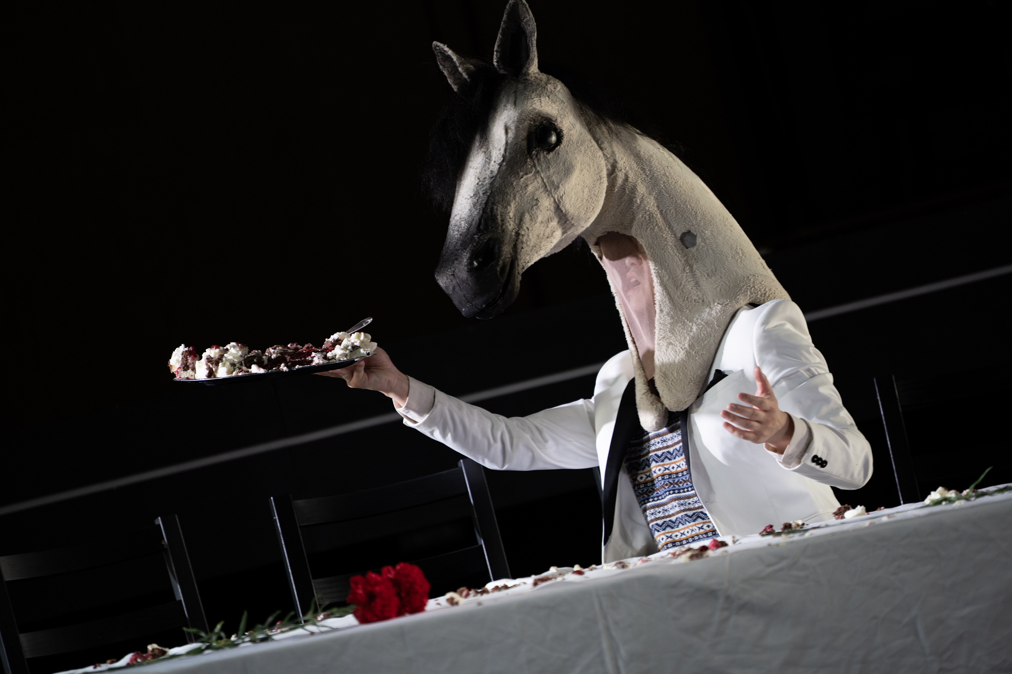 Eine Person mit weißem Pferdekopf hält einen Teller mit den Resten einer Sahnetorte in der Hand