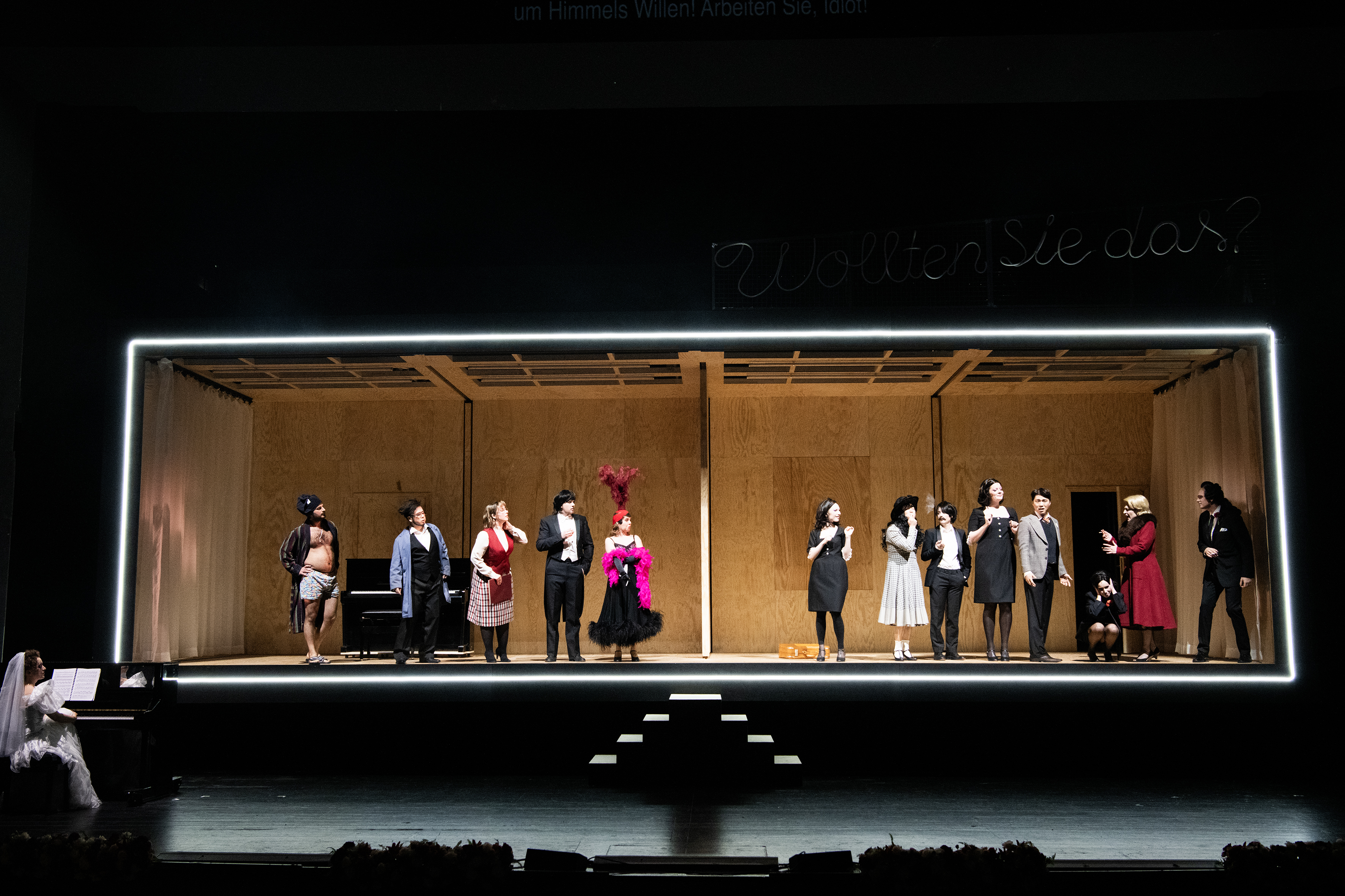 Dreizehn Darsteller:innen im Bühnenbild eines breiten Holzraums, die Umrandung ist erleuchtet
