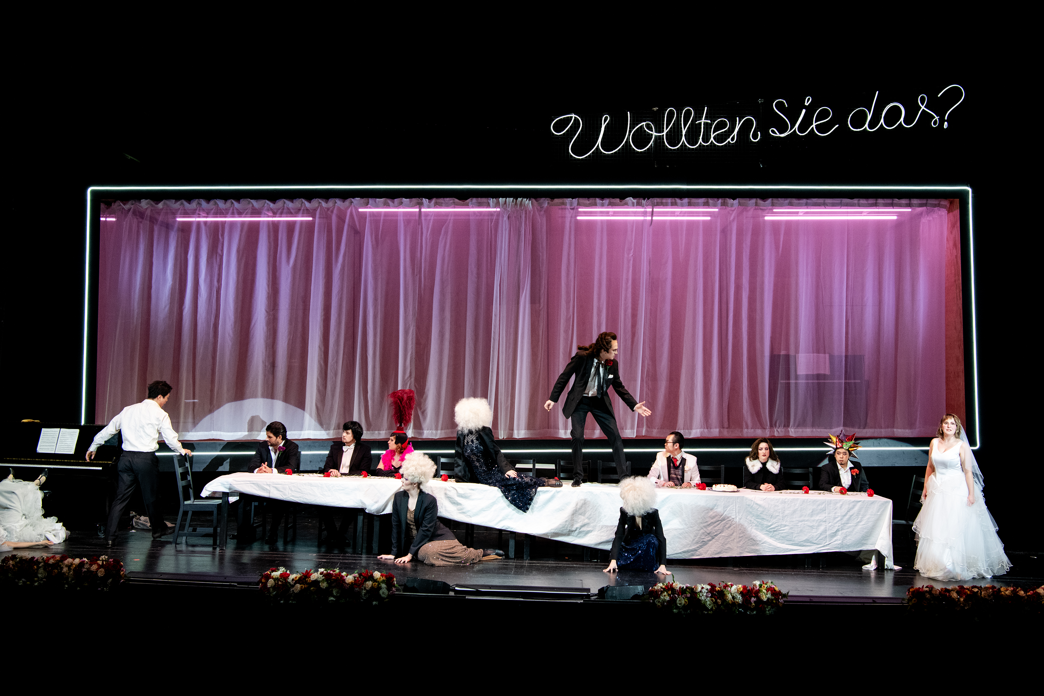 Eine gedeckte Tafel, an der Darsteller:innen sitzen oder davor knien. Eine Figur steht auf dem Tisch und blickt in Richtung einer Frau im Brautkleid neben dem Tisch