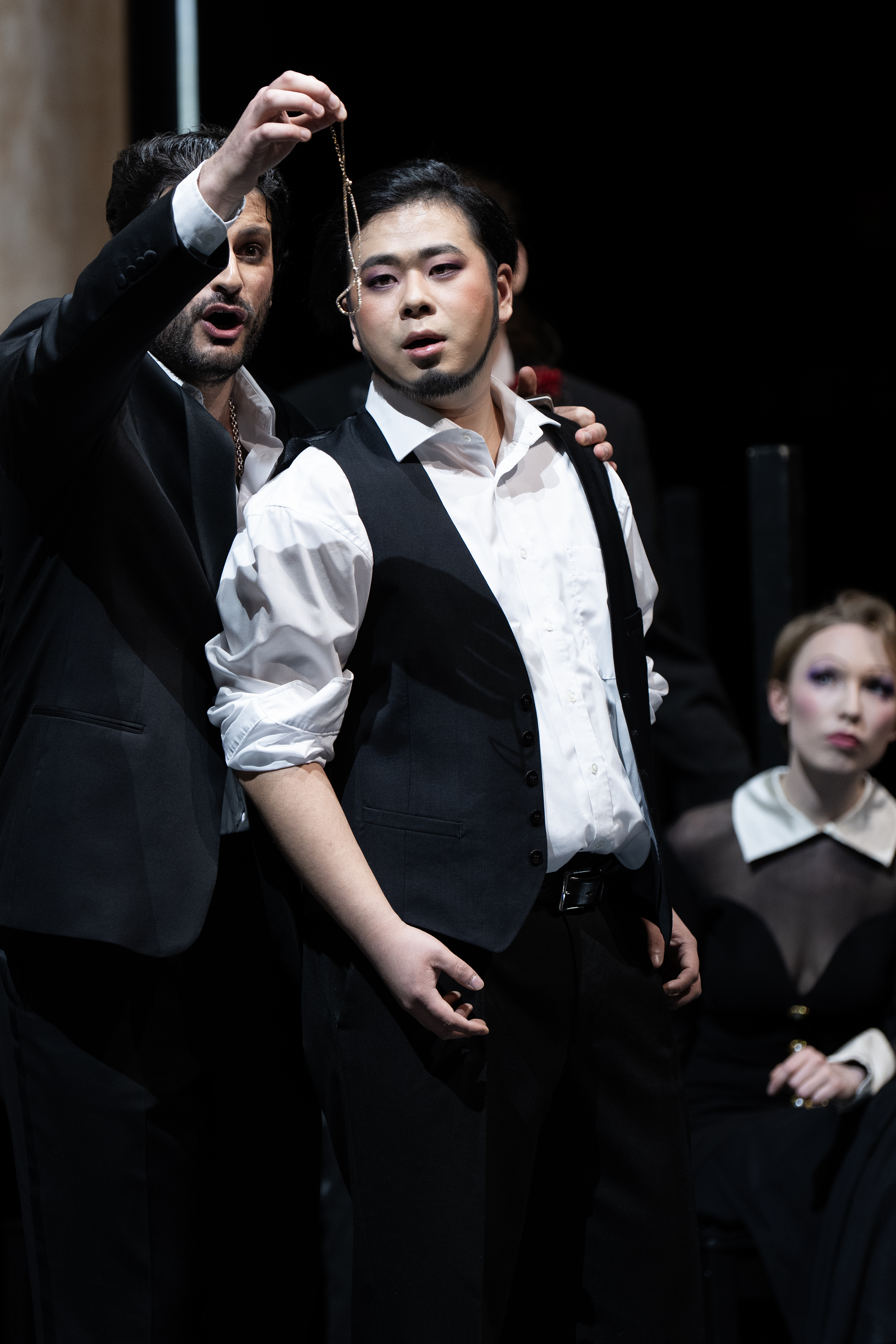 Zwei Männer, einer hält dem anderen eine Halskette vor sein Gesicht, im Hintergrund eine beobachtende Frau