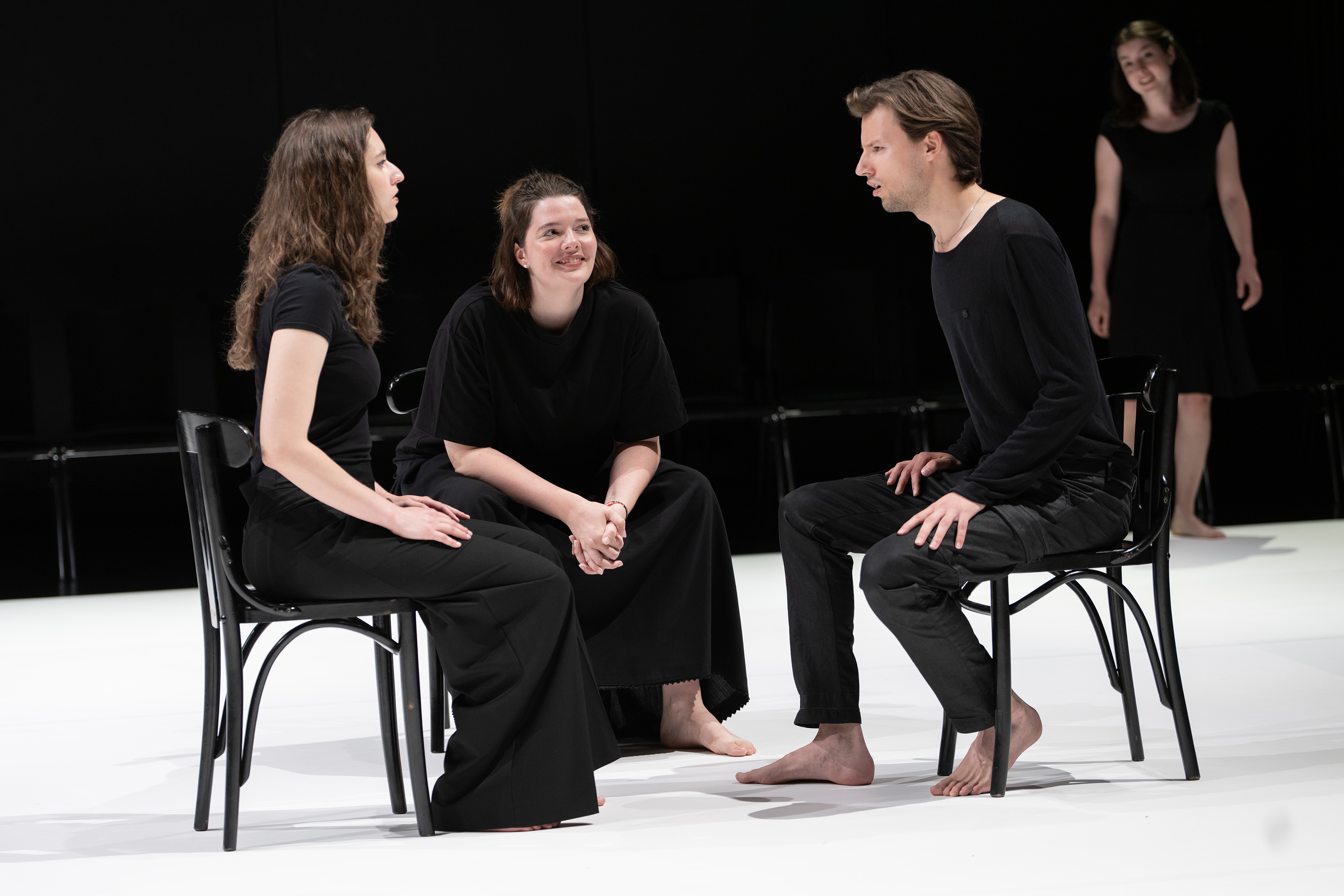 Szenenfoto: Drei Personen in schwarzer Kleidung (Katja Maderer, Viktoria Matt, Luis Weidlich) sitzen auf Stühlen und unterhalten sich, eine vierte Person (Julia Schneider) steht im Hintergrund