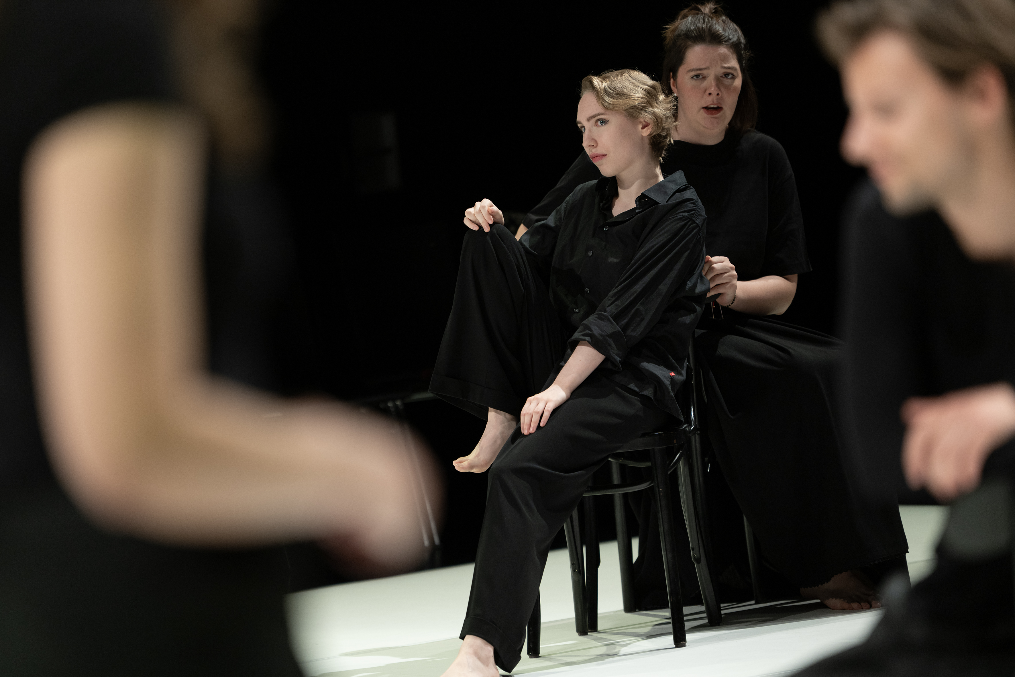 Szenenfoto: Zwei Frauen in schwarzer Kleidung (Madeleine Shari Wulff und Viktoria Matt) sitzen auf einem Stuhl vor schwarzem Hintergrund, im Vordergrund unscharfe Personenansichten