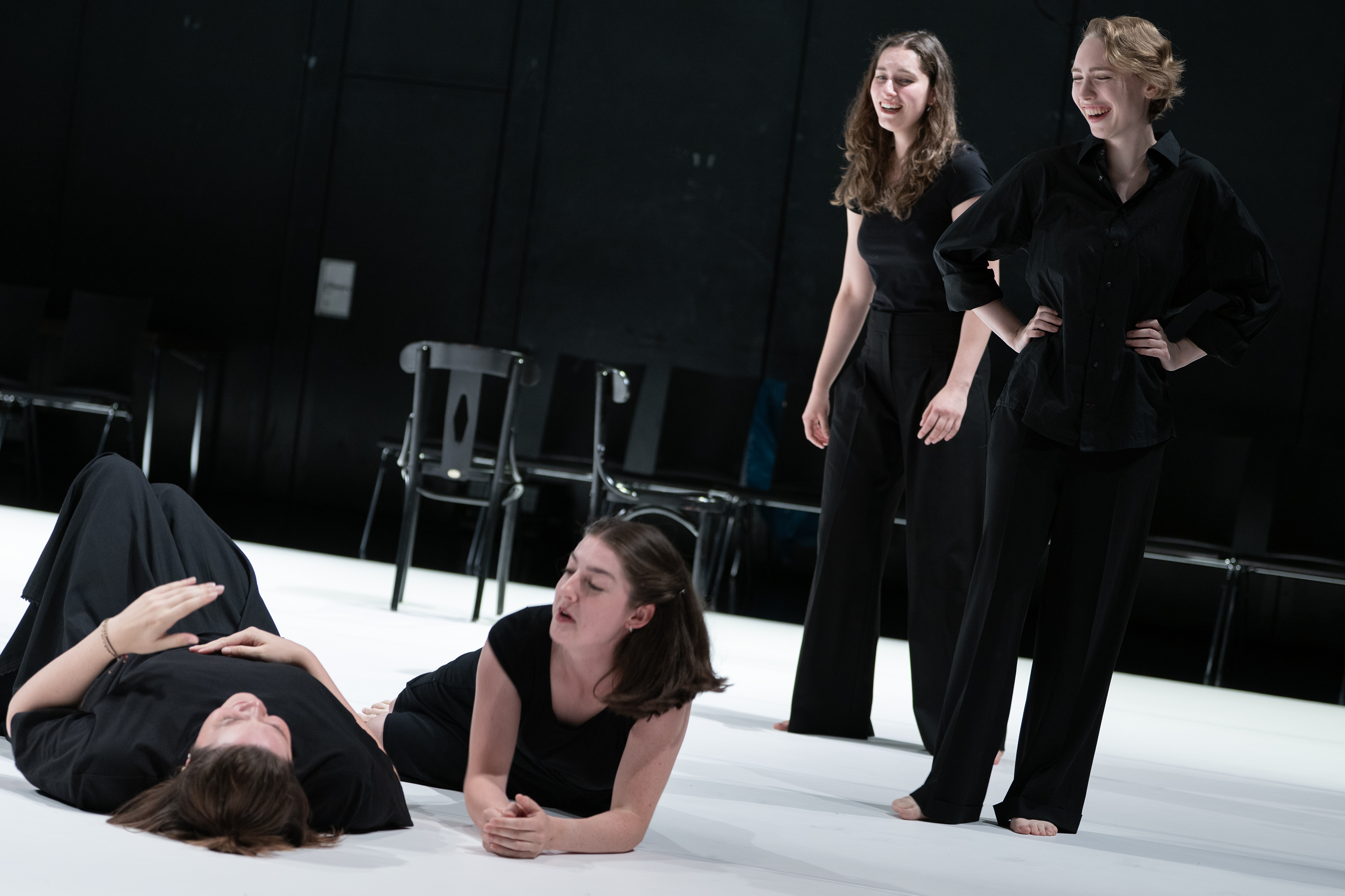 Szenenfoto: Vier Personen in schwarzer Kleidung auf einer hellen Bühne, zwei (Katja Maderer, Madeleine Shari Wulff) stehen lachend, zwei (Viktoria Matt, Julia Schneider) liegen auf dem Boden