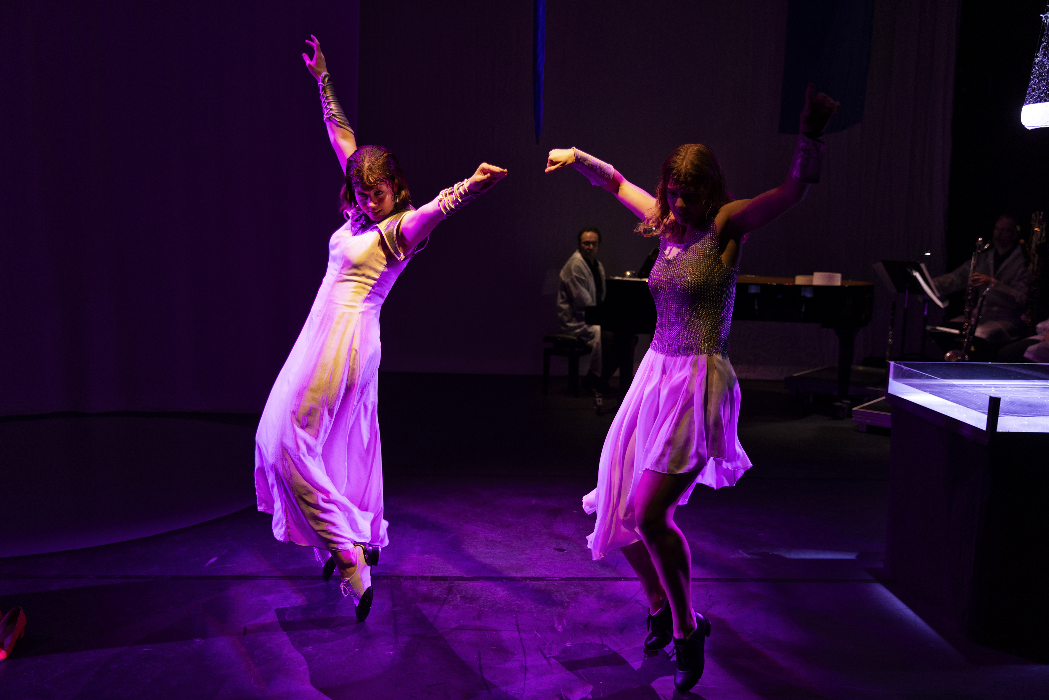 Szenenfoto: Zwei Frauen in langen Kleidern tanzen auf einer Bühne mit dunklem Hintergrund und violettem Licht, im Hintergrund sitzt ein Mann am Klavier
