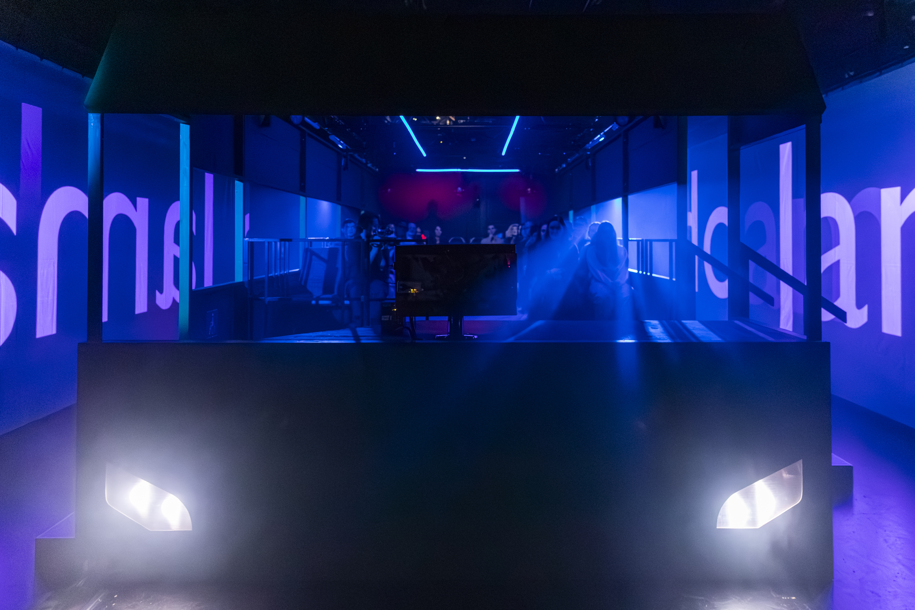 Das Bühnenbild in Form eines Buses, von vorne zu sehen, mit angeschalteten Scheinwerfern, links und rechts vom Bus Buchstaben in blauem Licht auf Leinwänden