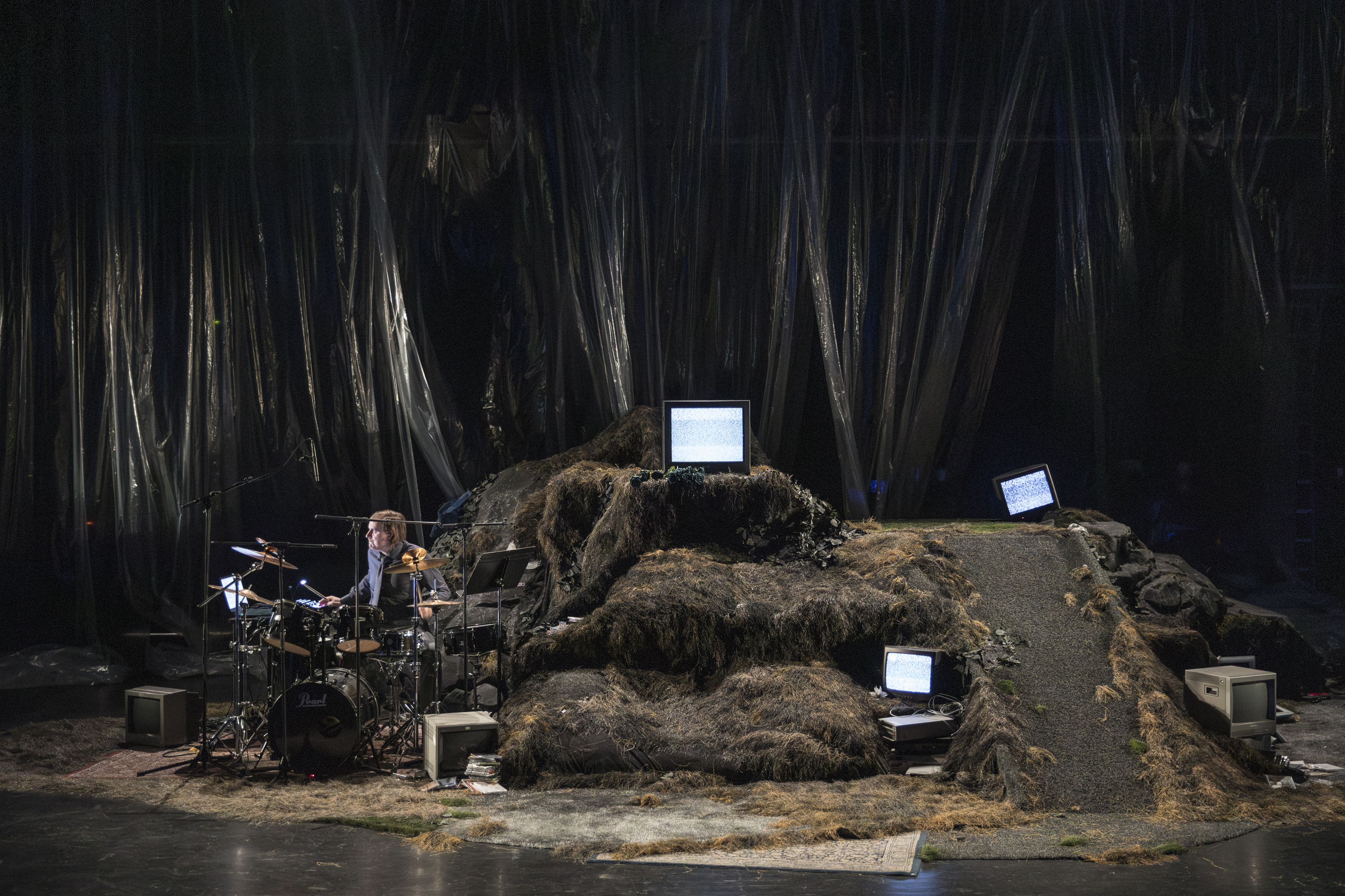 Das Bühnenbild bestehend aus Vorhängen aus Folie sowie analogen Fernsehern und Modems, auf der linken Seite ein Schlagzeug