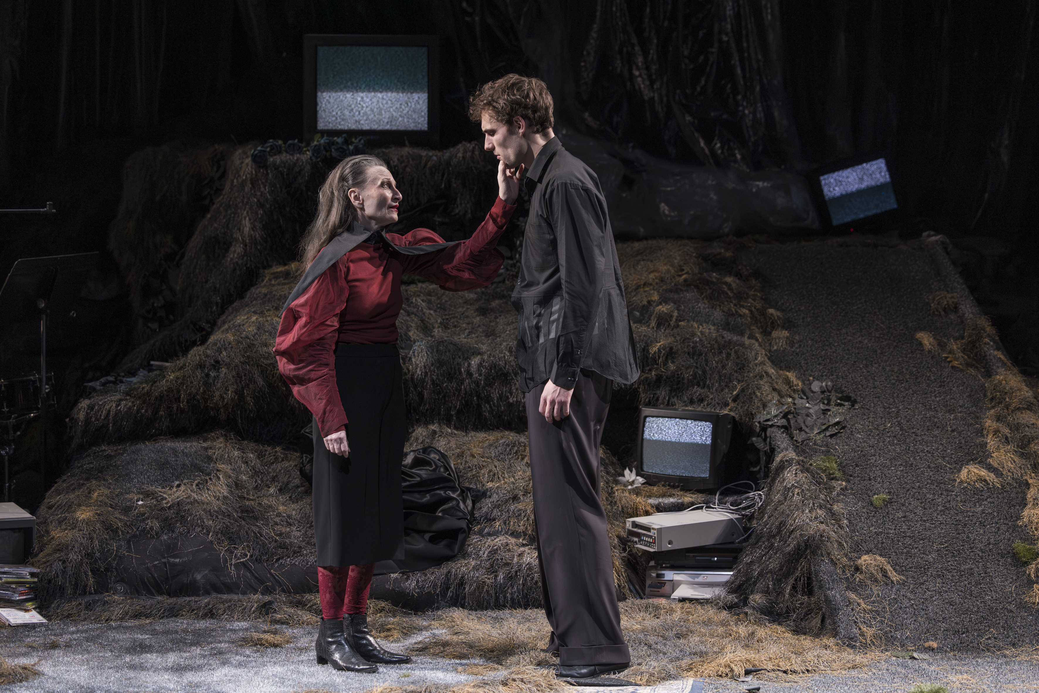 Eine ältere Frau steht vor einem jüngeren Mann, sie berührt mit ihrer linken Hand seine Wange