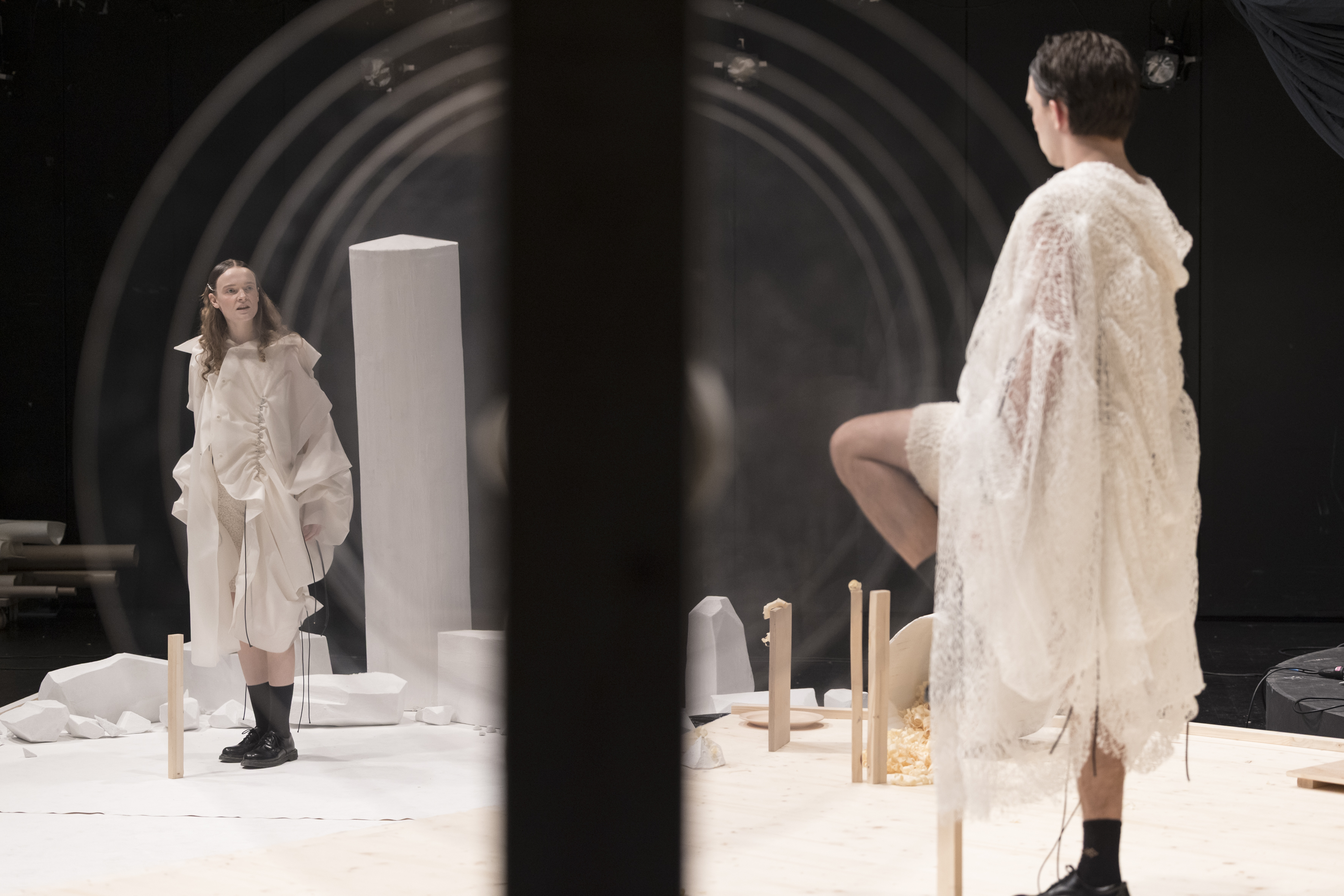 Zwei Personen in weißen, voluminösen Kleidern stehen auf einer Bühne mit weißen und hölzernen Requisiten vor schwarzem Hintergrund