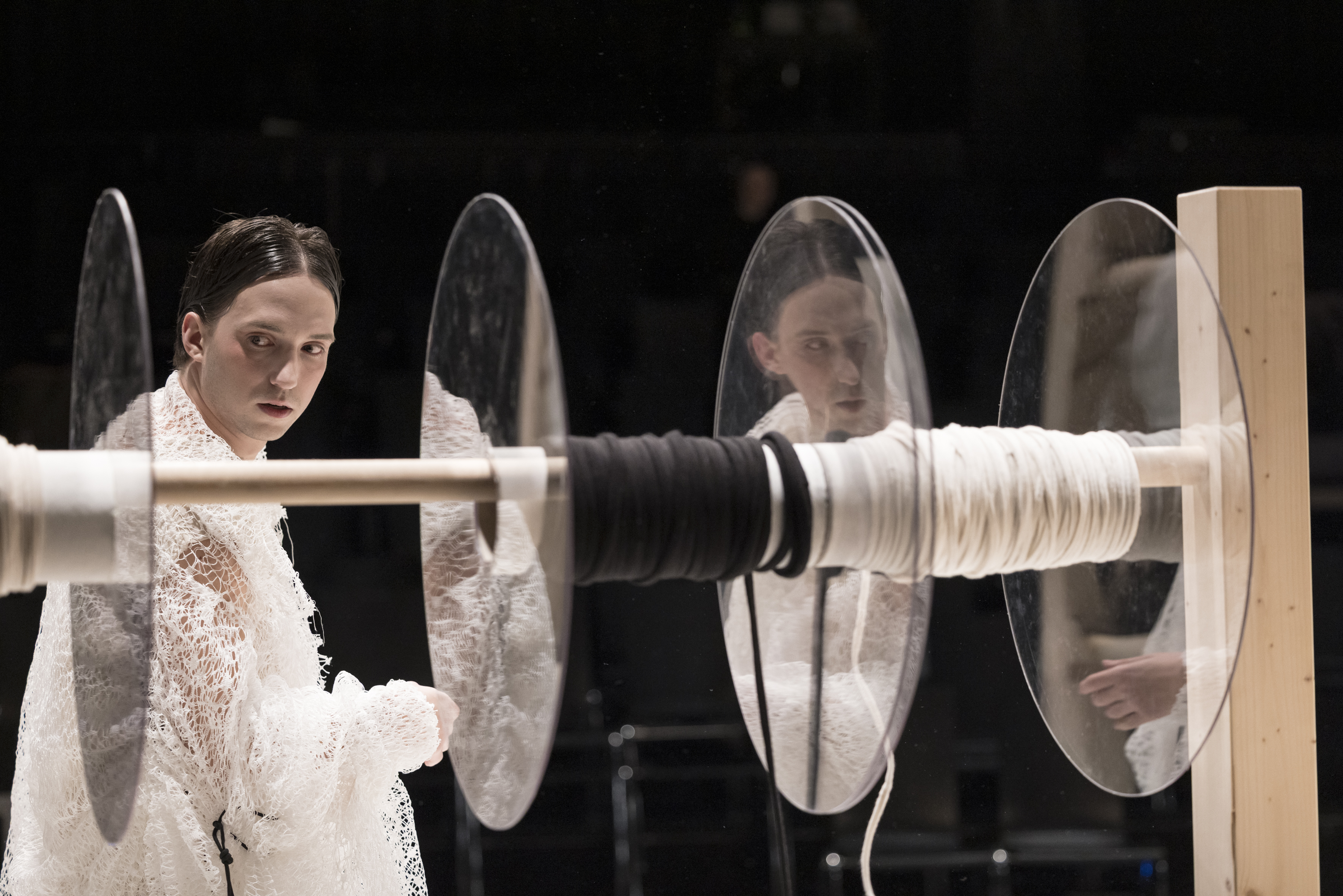 Ein Mann mit glatten schwarzen Haaren und weißem Gewand schaut nach links zurück, vor ihm eine horizontale Stange mit spiegelnden Glasscheiben und aufgewickelten schwarzen und weißen Seilen