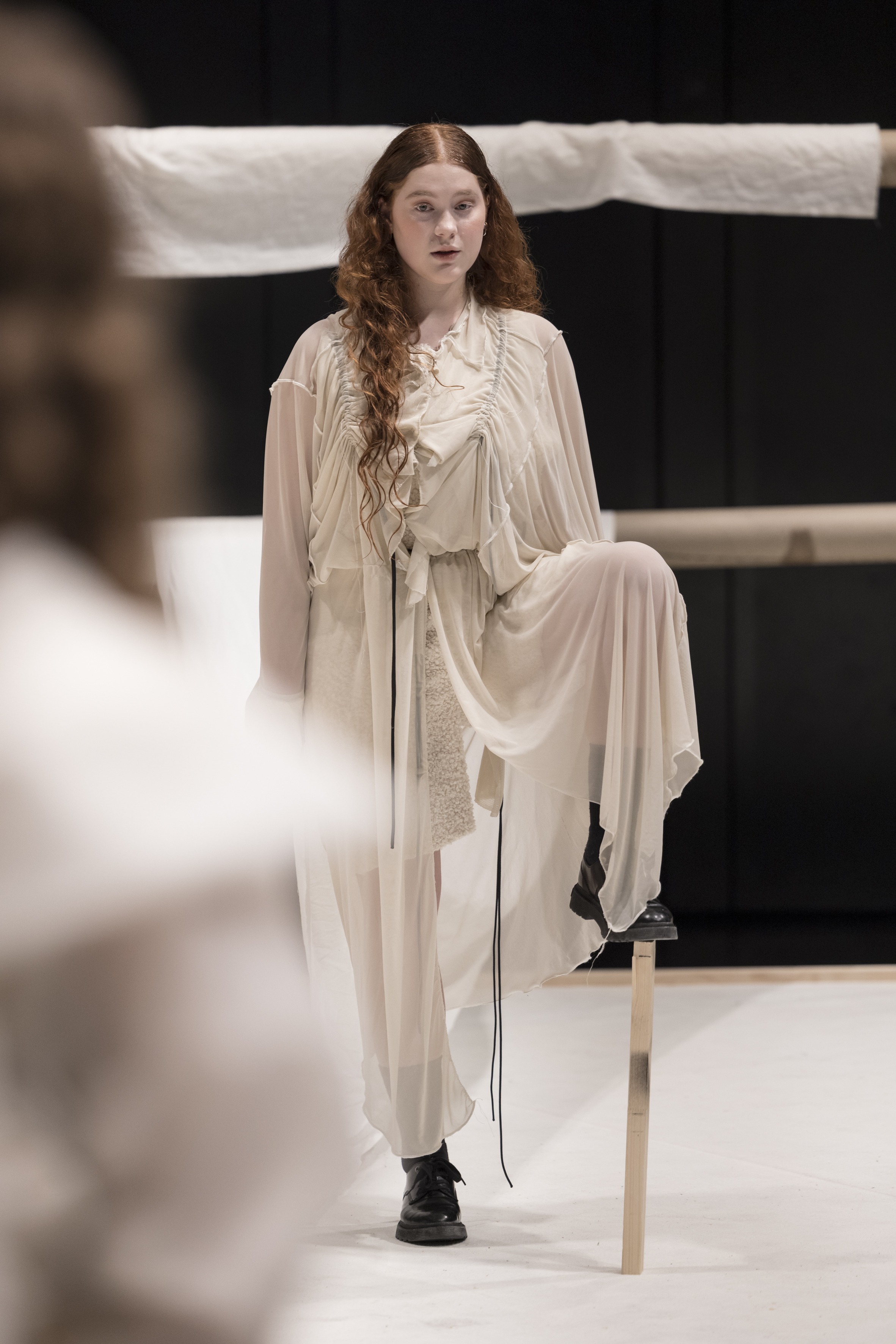 Person mit langen lockigen Haaren in fließendem, hellem Kleid und schwarzen Schuhen steht mit einem Bein auf einem Holzstab vor schwarzem Hintergrund