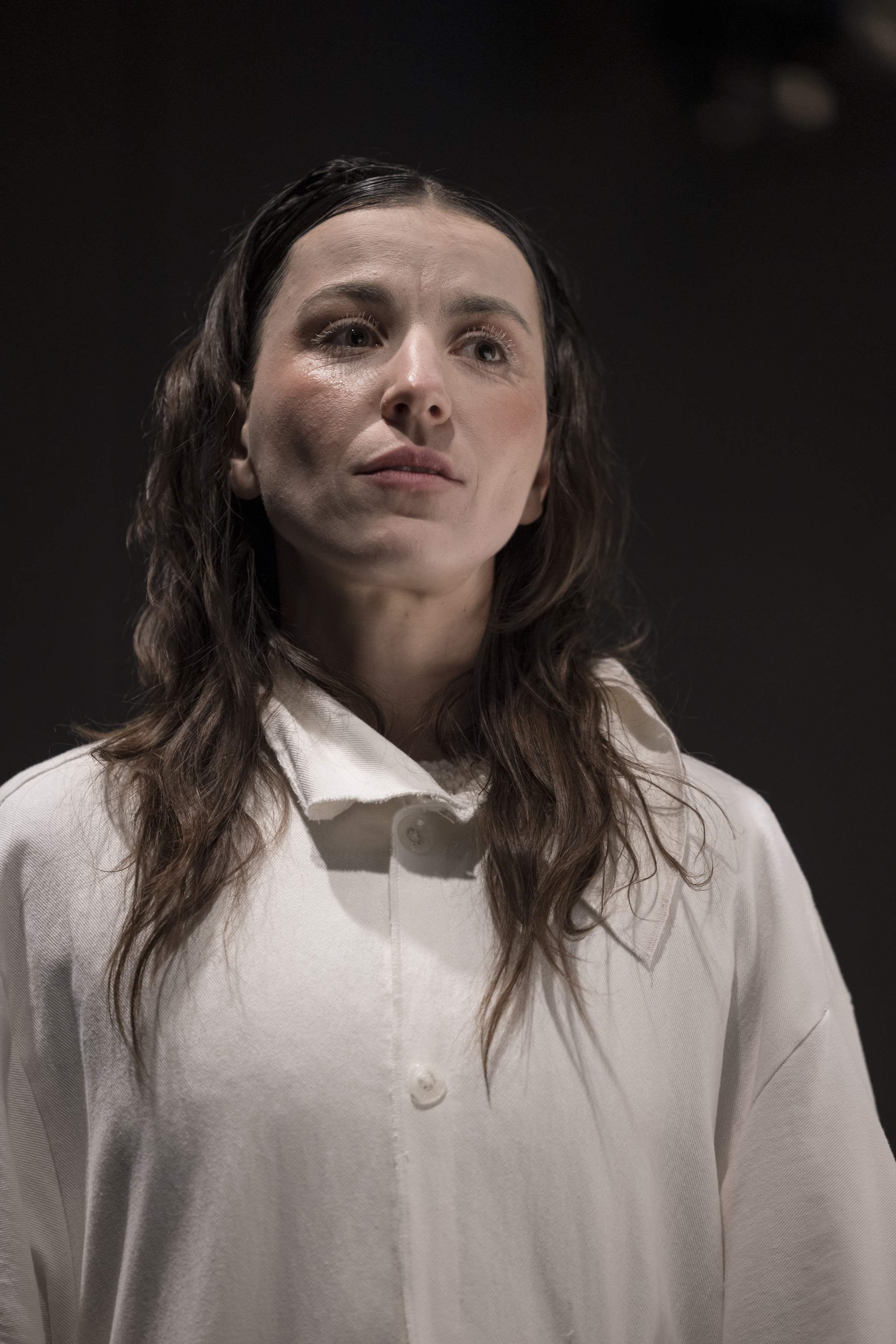 Person mit langen, gewellten Haaren trägt ein helles Hemd mit Knöpfen vor dunklem Hintergrund