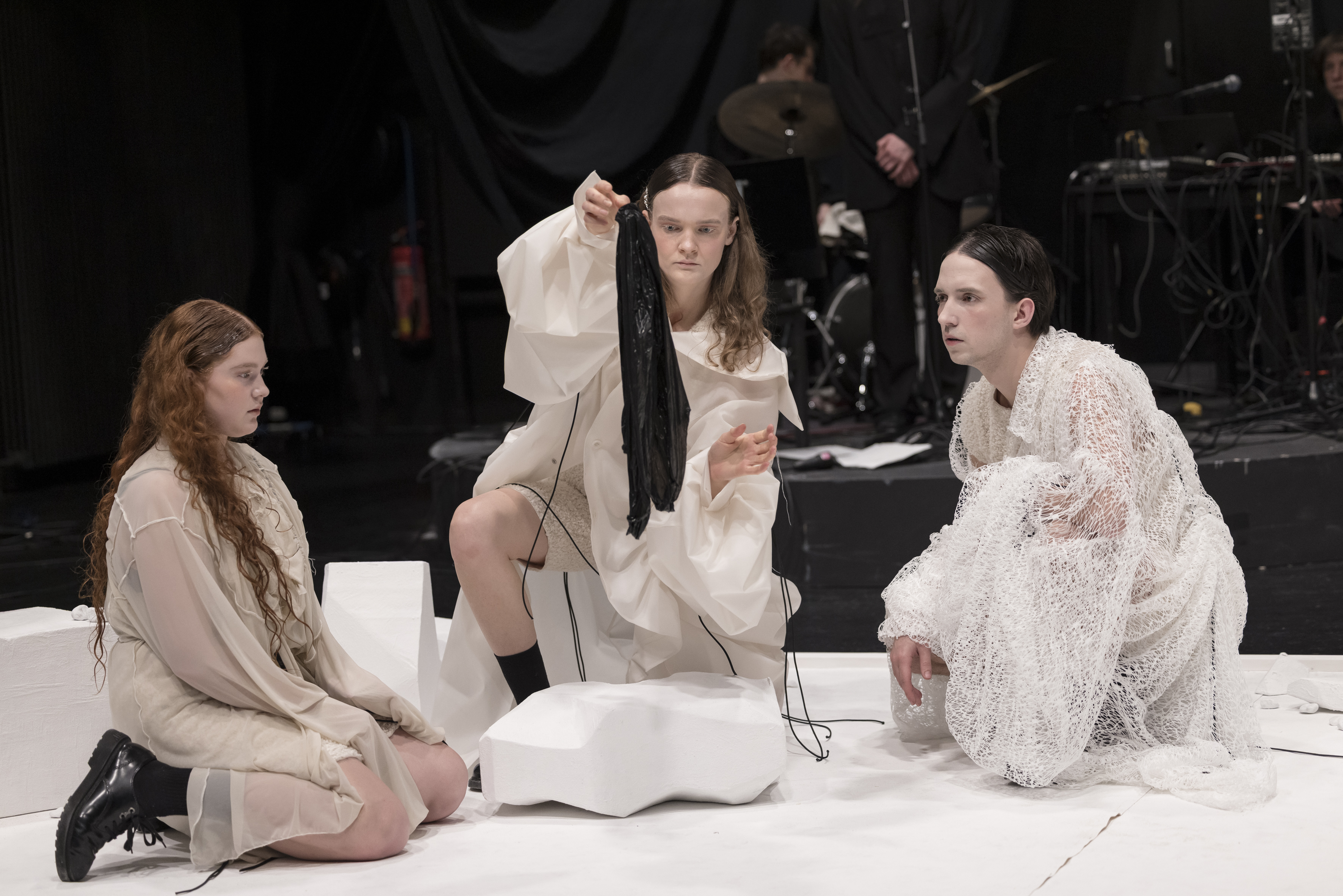 Drei Kaspare in weißen Gewändern kniend bzw. hockend auf einem weißen Boden, die Frau in der Mitte hält ein schwarzes Stück Stoff in die Luft, das alle ansehen