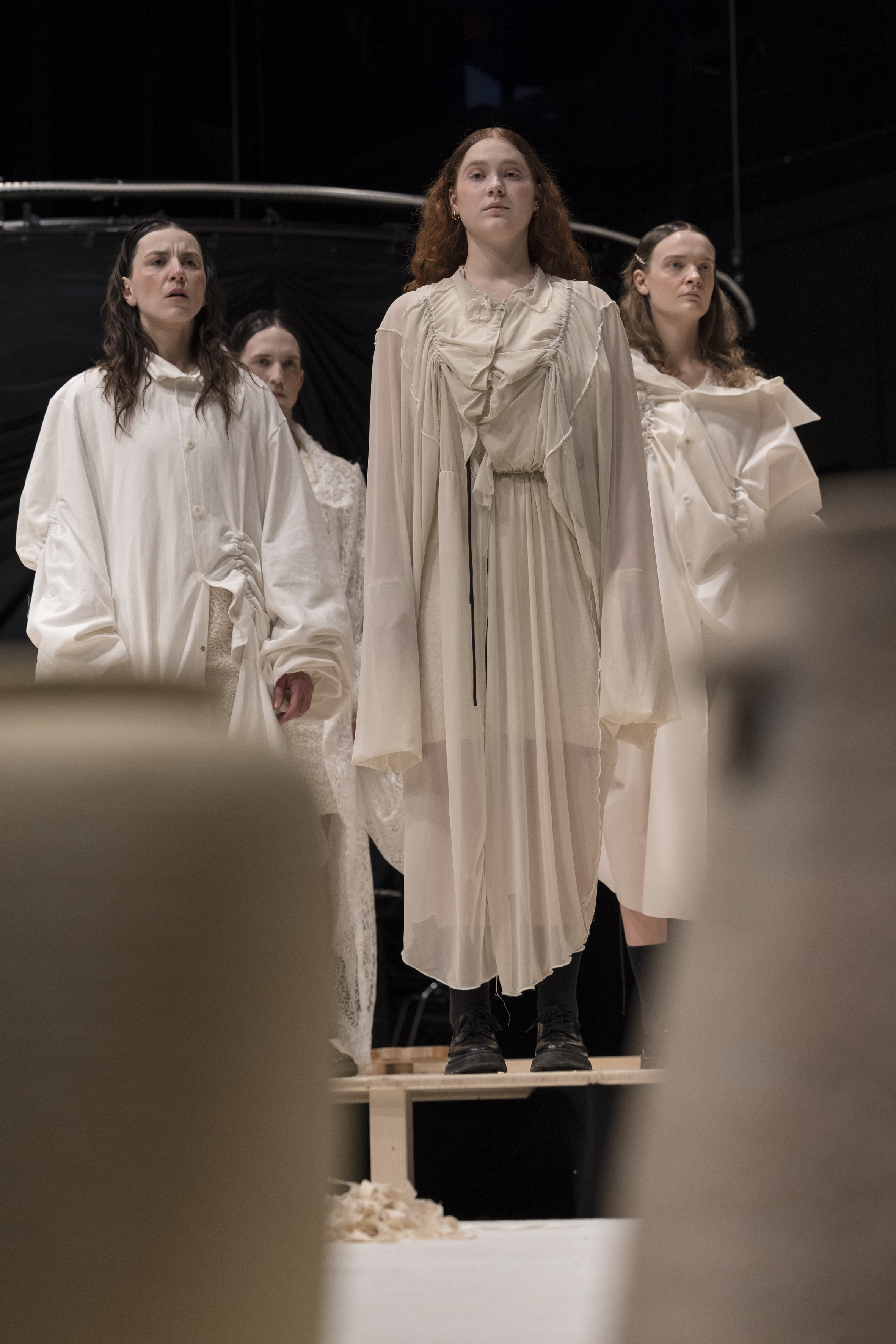 Vier Personen in weißen, fließenden Kleidern stehen auf einer Bühne, die vordere Person trägt schwarze Stiefel