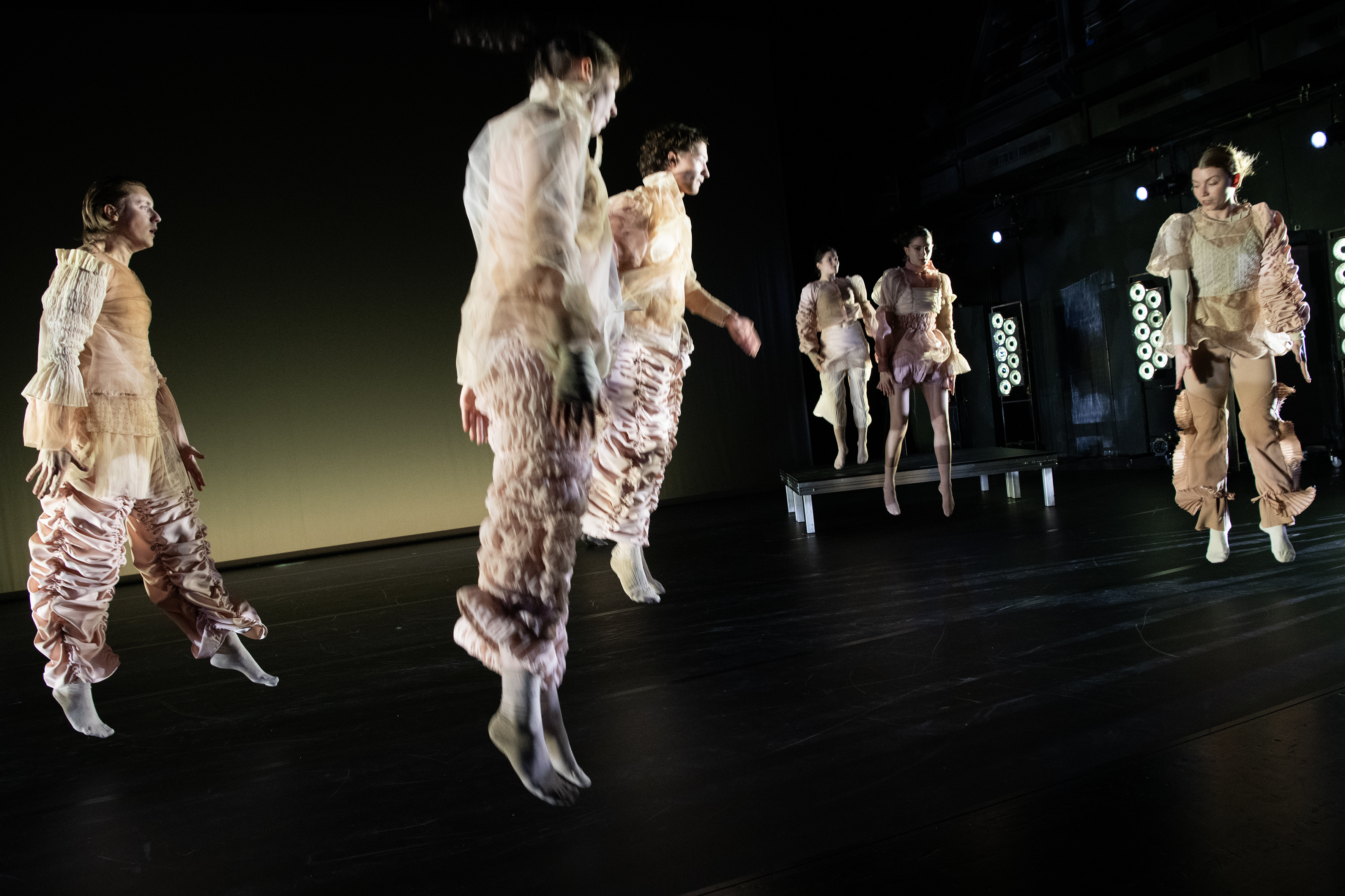 Ensemblemitglieder in hellen Kostümen in Bewegung oder im Sprung vor schwarzem Hintergrund