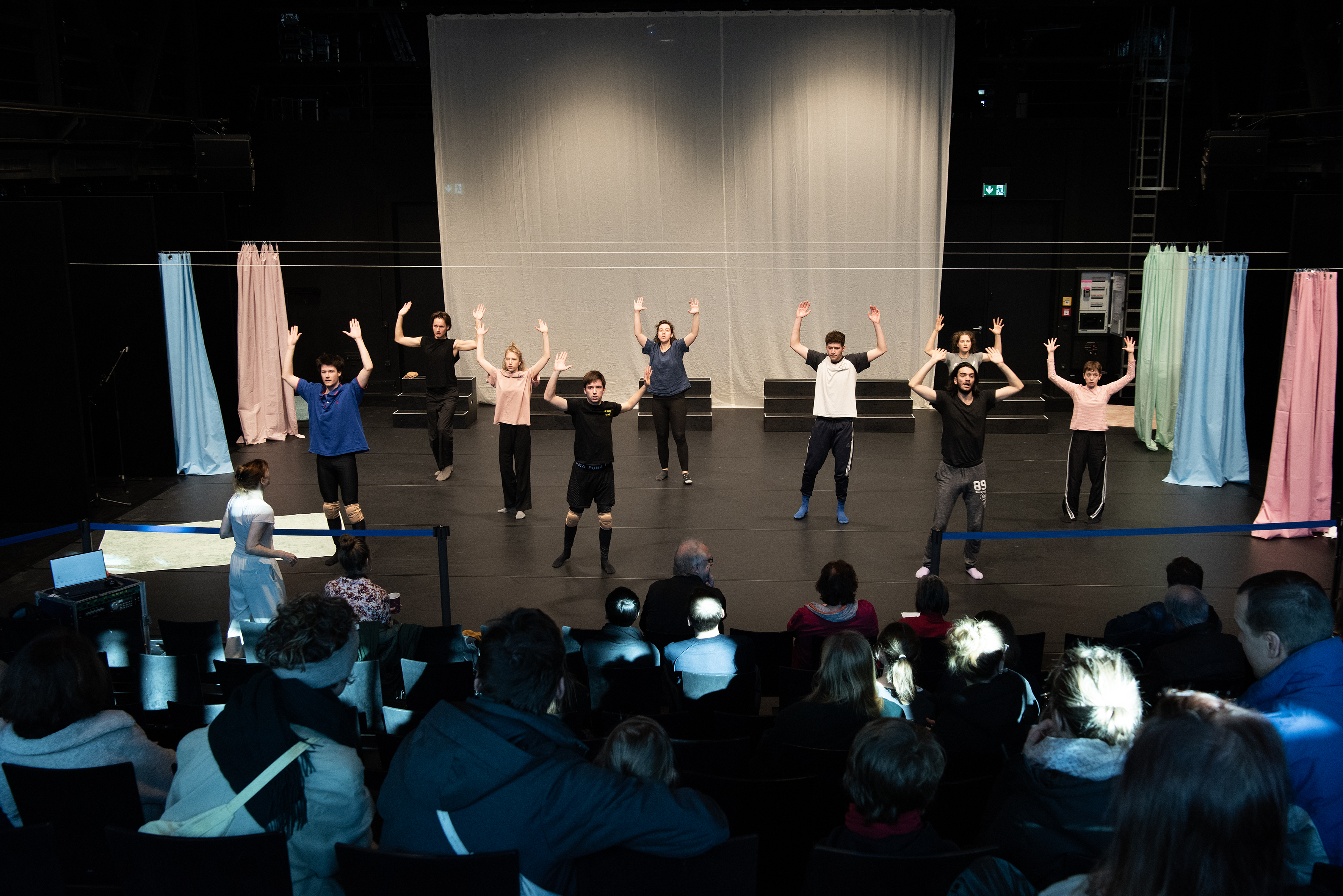 Auf einer Bühne stehen neun Schauspielstudierende mit erhobenen Armen vor Publikum