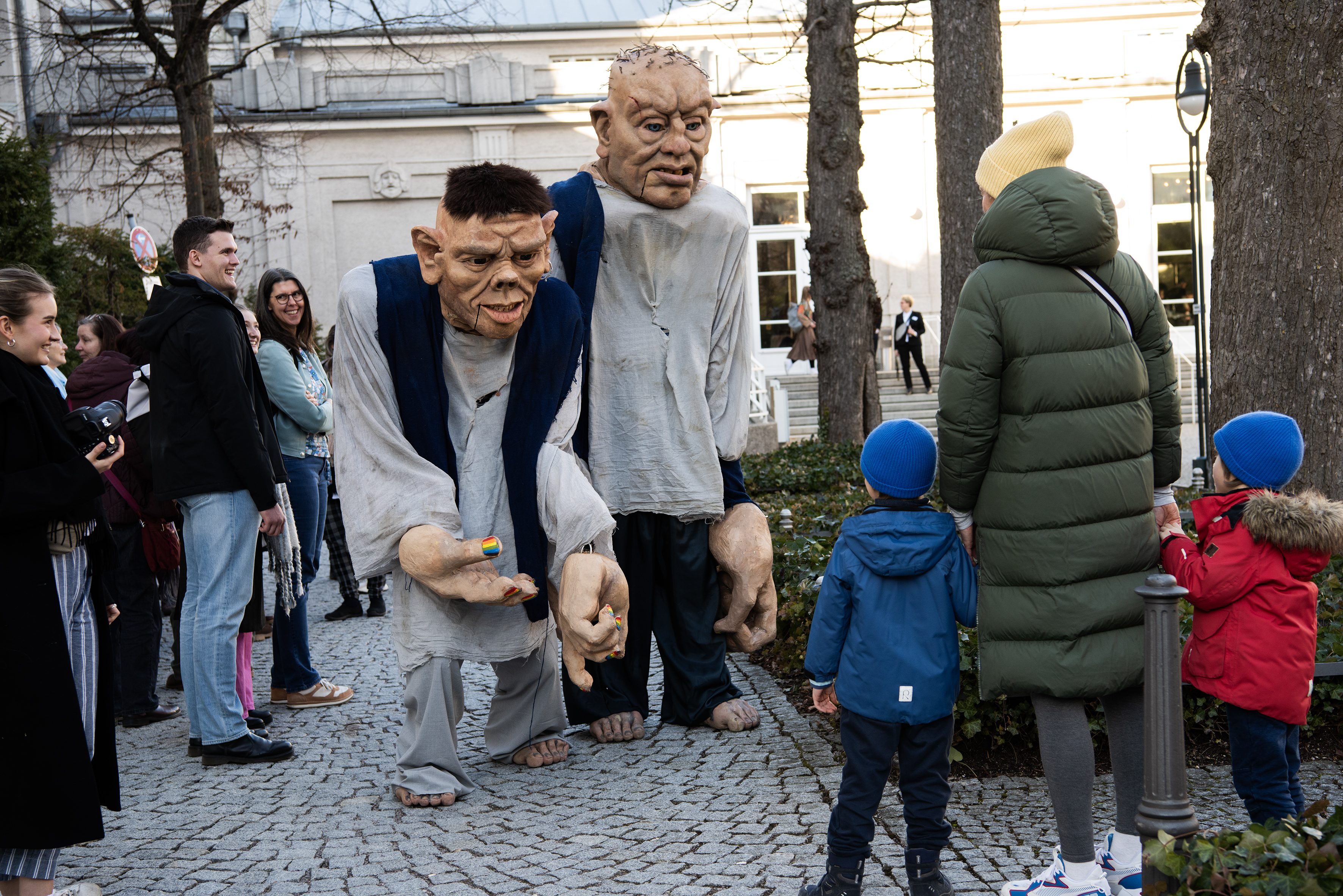 Zwei große Riesenfiguren stehen einer Frau und ihren zwei Kindern gegenüber und strecken eine Hand zur Begrüßung aus