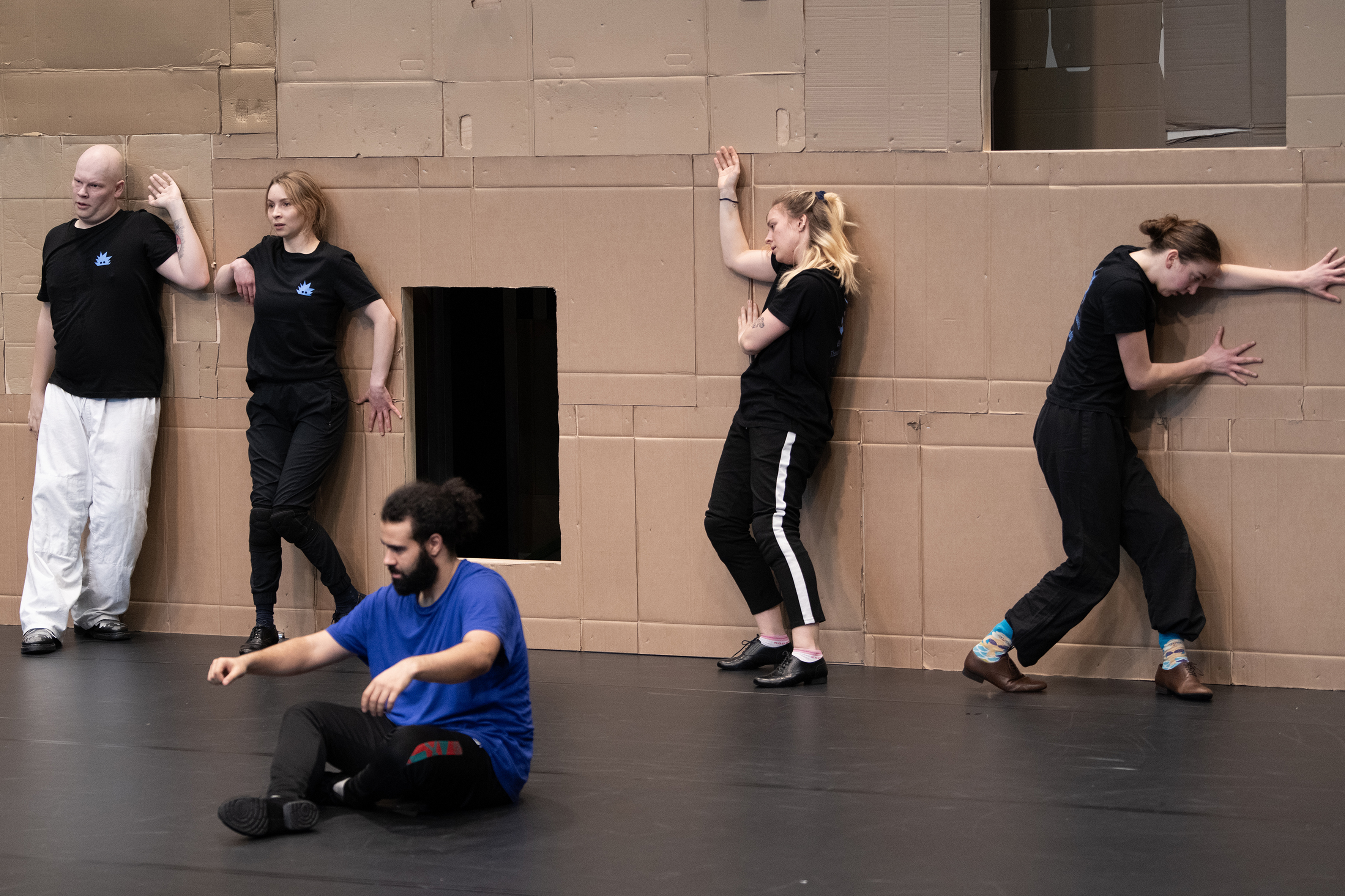 Vier Studierende stehen in verschiedenen Posen vor einer Wand, einer sitzt im Schneidersitz davor
