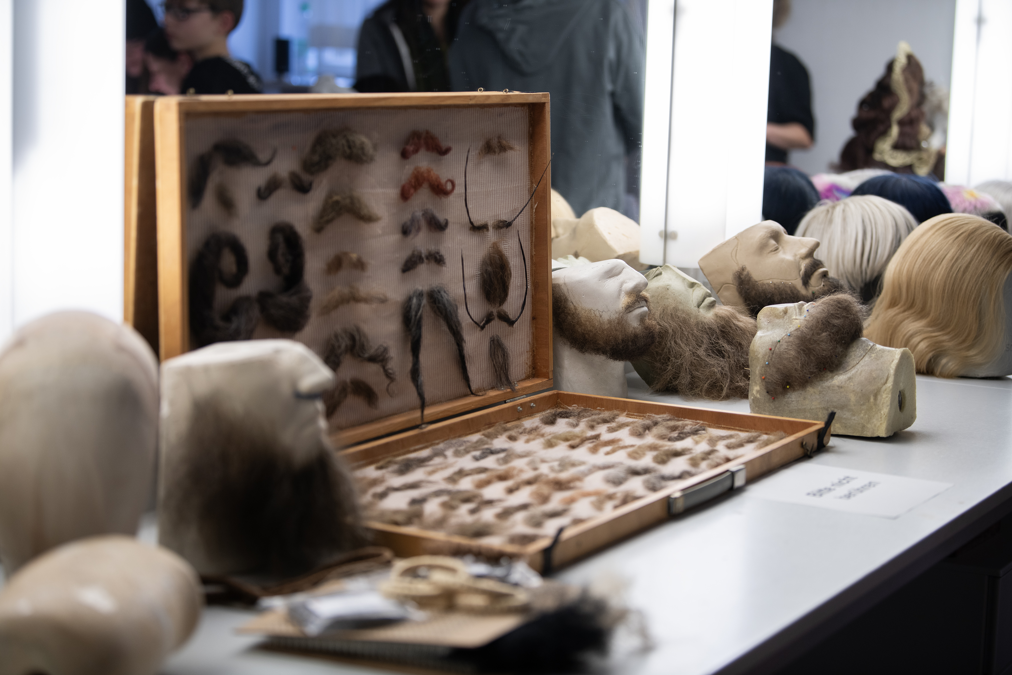 Ein geöffneter Kasten mit Schnurrbärten, umgeben von Köpfen und Haarteilen vor einem Maskenbildspiegel
