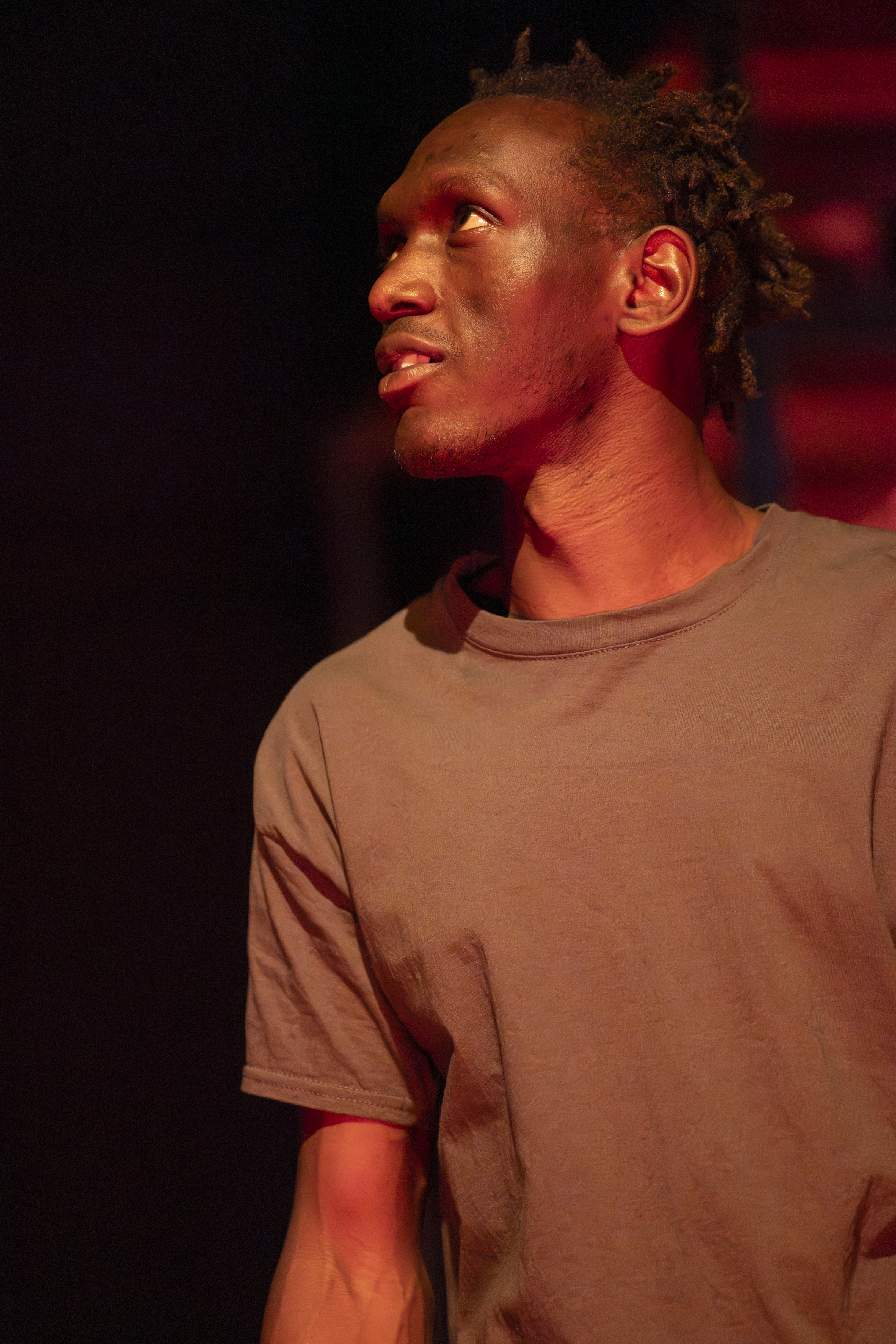 Szenenfoto: Ein Schwarz gelesener Mann (Duncan Lubwama) mit kurzen Haaren trägt ein schlichtes T-Shirt und steht vor einem dunklen, rot beleuchteten Hintergrund