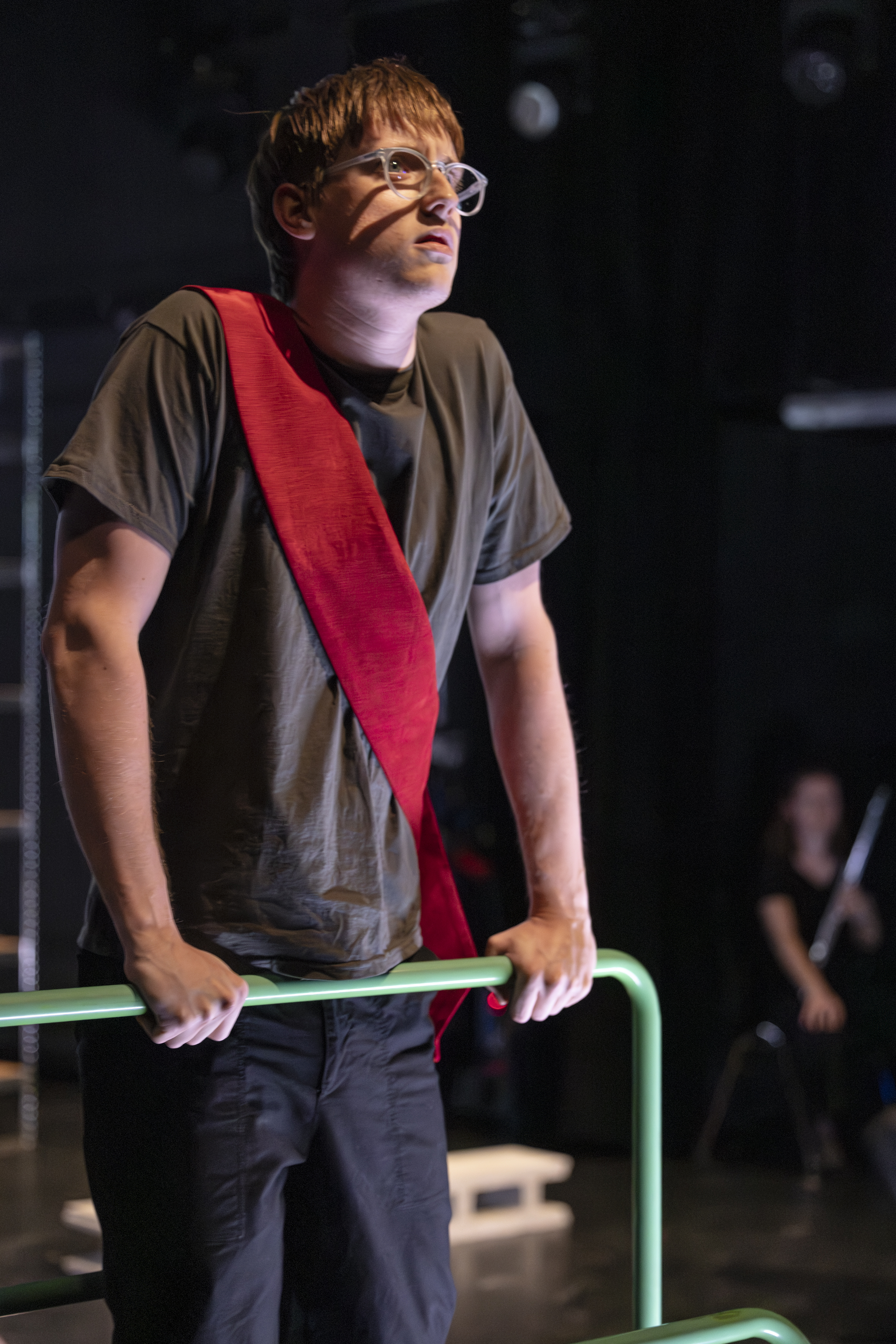 Szenenfoto: Ein weiß gelesener Mann (David Jakob Hirmer) mit Brille, rotem diagonalen Stoffband über der Schulter, grauem T-Shirt und dunkler Hose lehnt an grünem Geländer in dunklem Raum