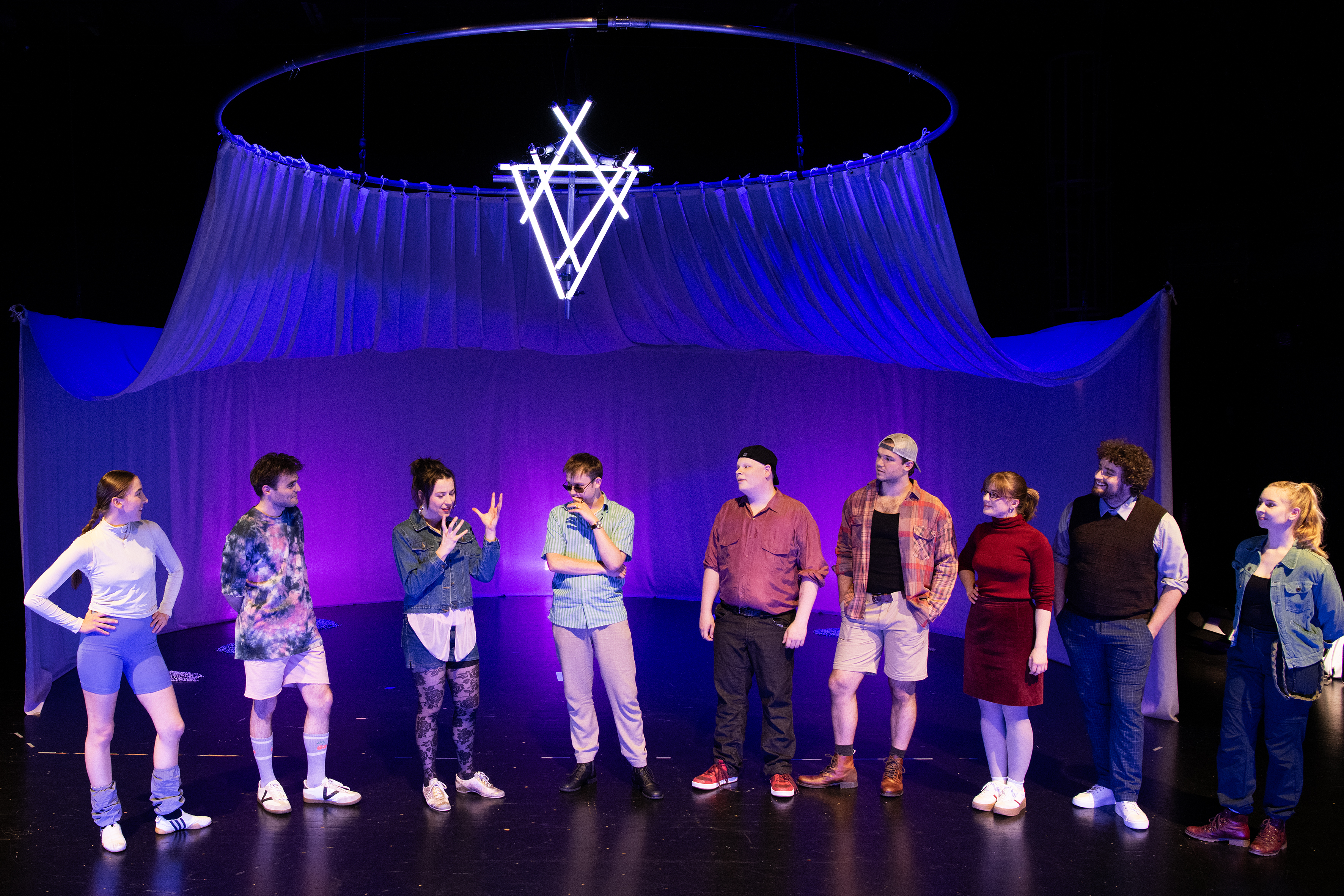 Neun Personen in bunter Kleidung stehen auf einer Bühne vor einem violetten Hintergrund mit einem leuchtenden geometrischen Symbol darüber