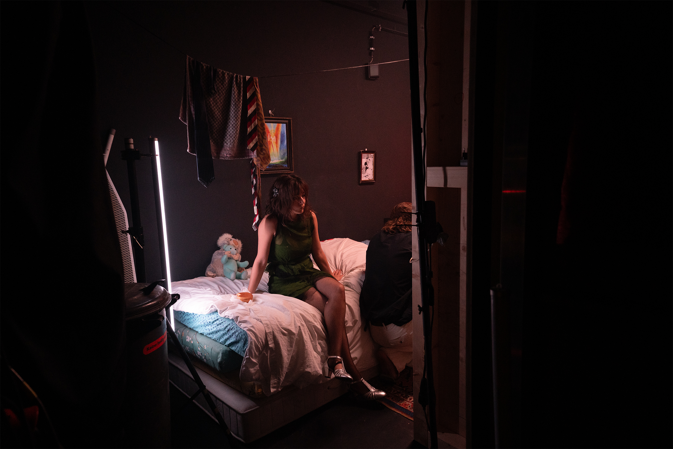 Szenenfoto: Frau sitzt auf einem Bett in einem dunklen Raum mit einem stehenden Licht und einem Kuscheltier im Hintergrund